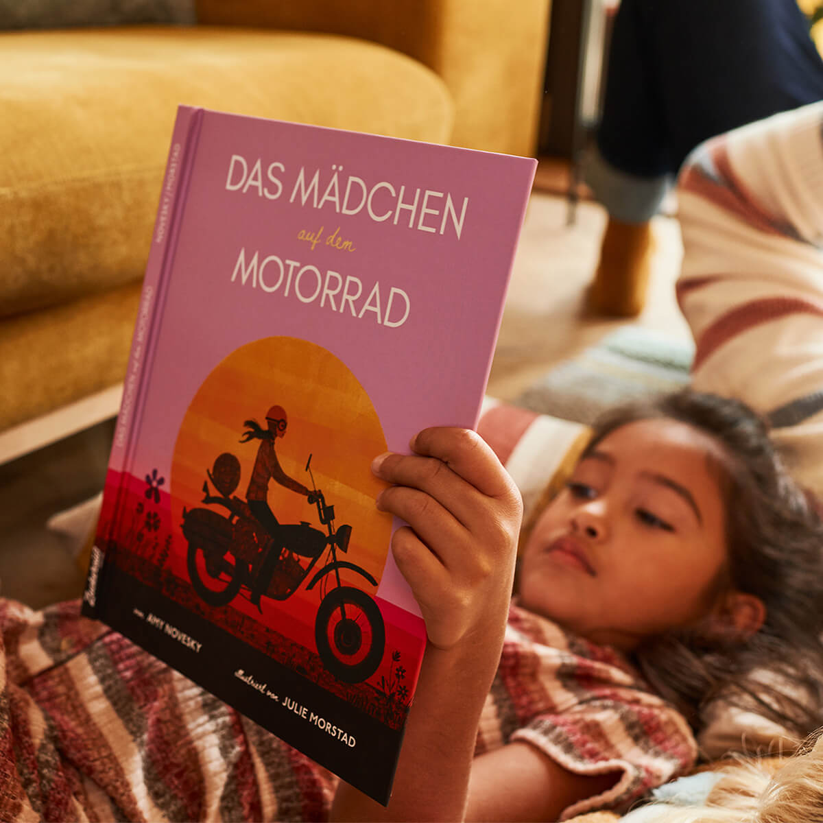 Ein kleines Kind liest "Das Mädchen auf dem Motorrad" von Amy Novesky, ein biografisches Bilderbuch über Anne-France Dautheville, das ein illustriertes Mädchen auf einem Motorrad bei Sonnenuntergang zeigt. Im Hintergrund erscheinen ein gelbes Sofa und eine weitere Person.