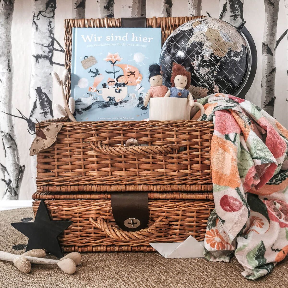 Das Kinderbuch „Wir sind hier“ von Kyo Maclear steht auf zwei geflochtenen Weidenkoffern, umgeben von einem schwarzen Globus, zwei Stoffpuppen in einer Tasse, einem bunten Handtuch, Holzspielzeug und einem Papierboot – eine liebevolle Szene, die Heimat und Zusammenhalt symbolisiert.
