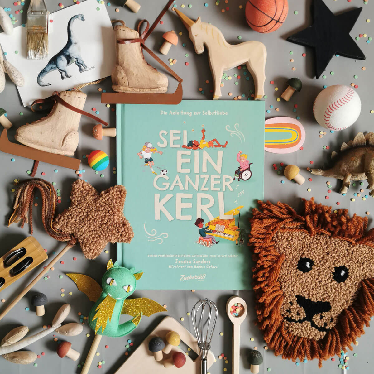 Das Kinderbuch „Sei ein ganzer Kerl“ liegt mittig auf einem grauen Untergrund, umgeben von buntem Konfetti, Holzspielzeug, Schlittschuhe, Einhorn, Dinosaurier, Stofffiguren (Löwe, Drache), Sportbällen, Kochutensilien und Bastelmaterialien.