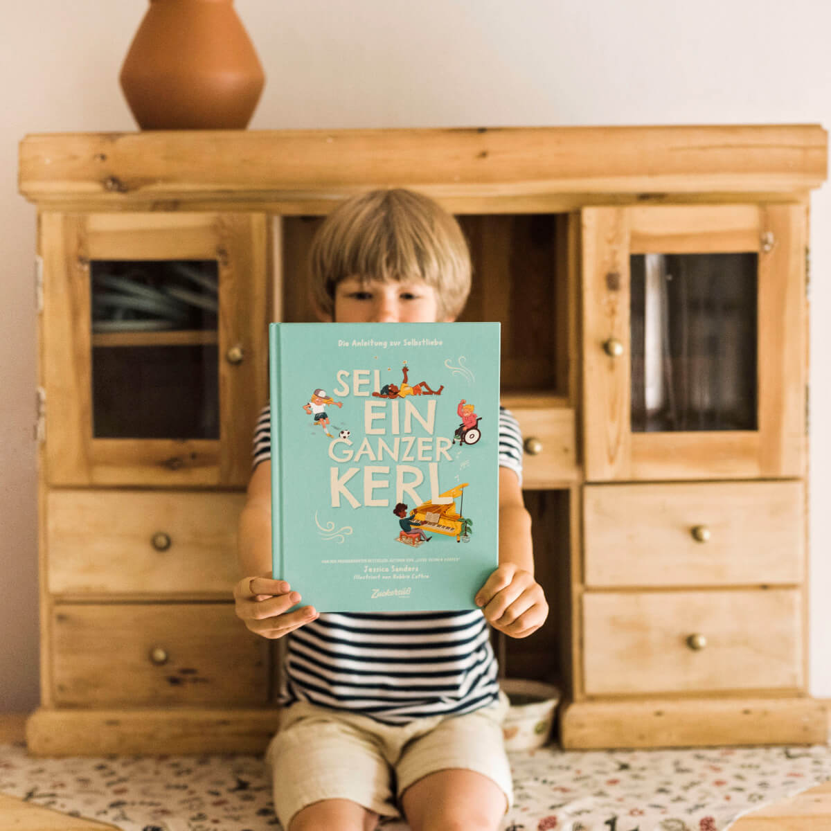 Ein blondes Kind in gestreiftem T-Shirt und Shorts sitzt auf einem gemusterten Teppich auf dem Boden vor einem rustikalen Holzschrank und hält das Buch „Sei ein ganzer Kerl“ von Jessica Sanders mit ausgestreckten Armen hoch, sodass das Gesicht bis auf die Augen fast vollständig verdeckt ist.
