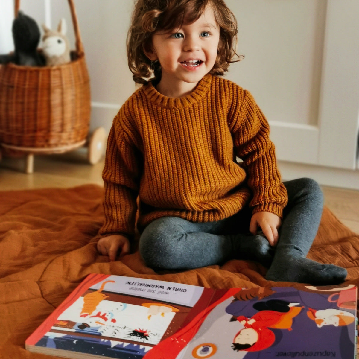 Ein kleines Kind mit lockigen braunen Haaren, das Susann Hoffmanns "Kleidung ist für alle da!" - einen senfgelben Pullover und graue Leggings - trägt, sitzt lächelnd auf einer braunen Decke im Haus, ein aufgeschlagenes Kinderbuch vor sich, um das Selbstvertrauen zu stärken.