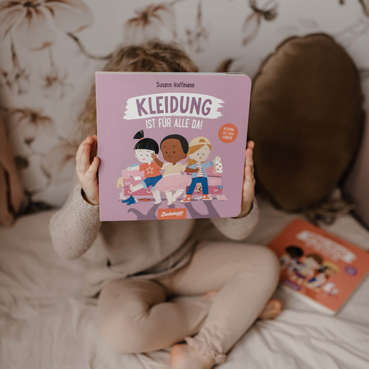 Ein Kind sitzt in beigefarbener Kleidung auf einem Bett mit floral gemusterter Tapete und hält das Buch „Kleidung ist für alle da!“ von Susann Hoffmann vor das Gesicht. Auf dem Cover sind drei gezeichnete Kinder in verschiedenen Kleidungsstilen zu sehen. Neben dem Kind liegt ein weiteres Buch aus derselben Reihe auf dem Bett.
