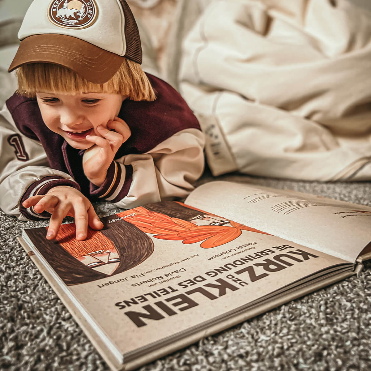 Ein kleines Kind mit Mütze und Jacke zeigt lächelnd auf ein aufgeschlagenes Bilderbuch, "Kurz und Klein" von Alastair Chisholm, das Geschichten über Höhlenmenschen und die Kraft der Zusammenarbeit erzählt.