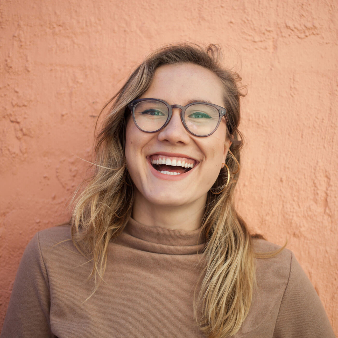 Jessica Sanders, die Autorin von „Liebe deinen Körper“, lächelt mit langen blonden Haaren und Brille vor einer pfirsichfarbenen Wand. In einem hellbraunen Rollkragenpullover strahlt sie Selbstliebe aus.