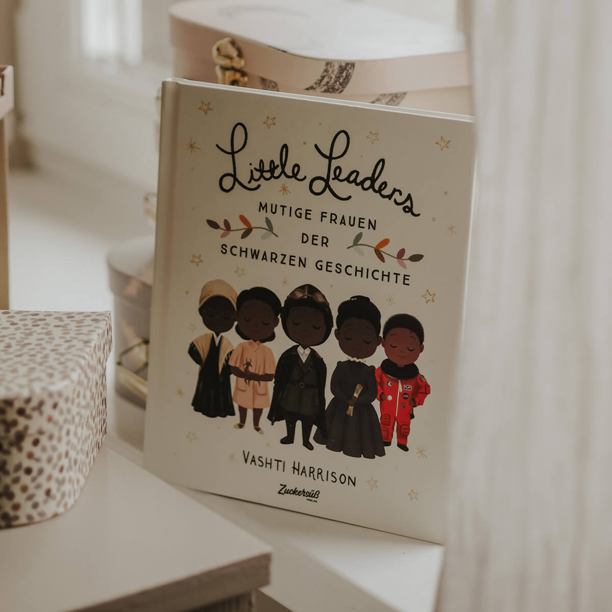 Ein Exemplar von „Little Leaders“ von Vashti Harrison steht aufrecht auf einem Schreibtisch, umgeben von dekorativen Boxen. Das Cover zeigt Illustrationen von fünf schwarzen Frauen.