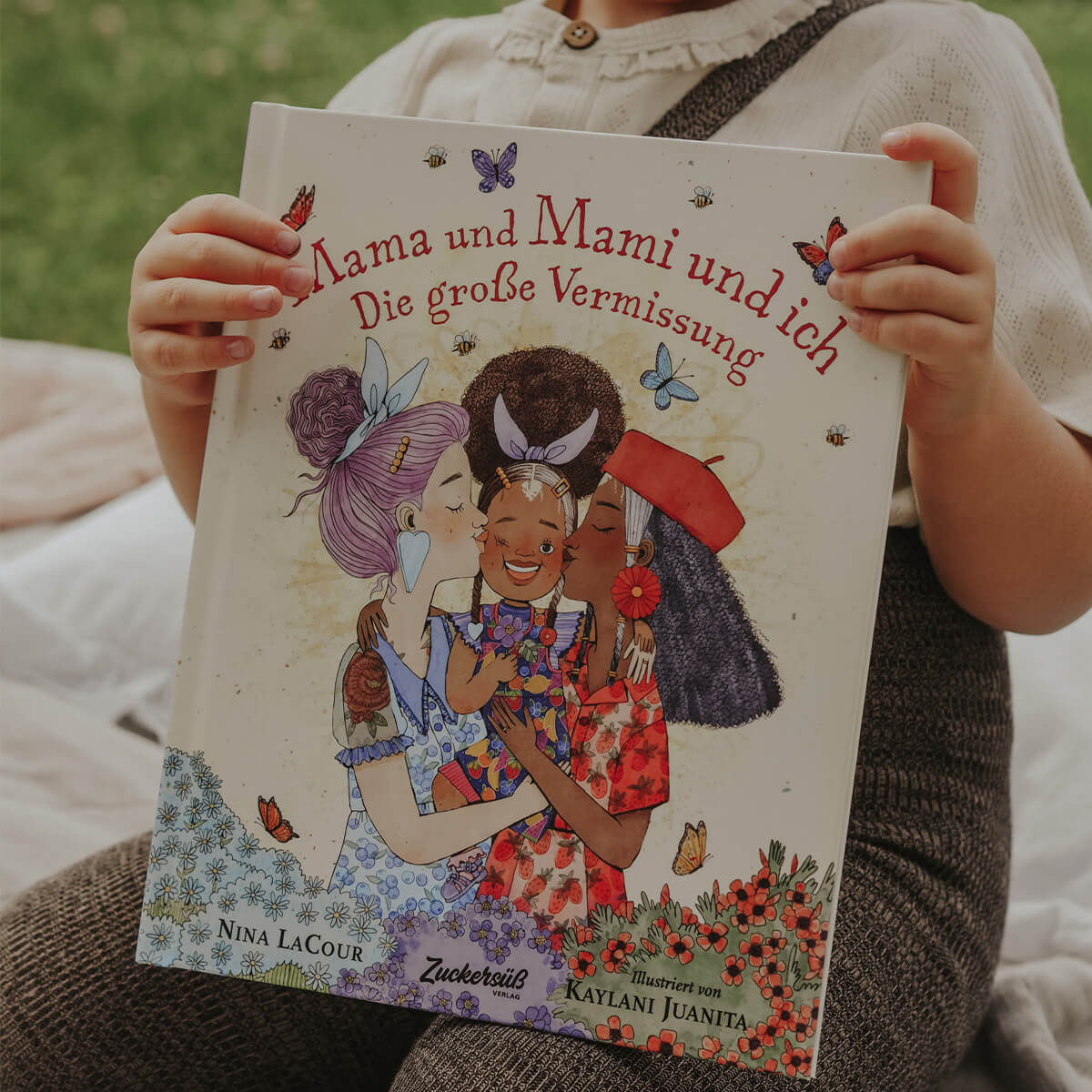 Ein Kind hält das Bilderbuch „Mama und Mami und ich: Die große Vermissung“ von Nina LaCour in den Händen. Auf dem Cover wird ein Kind liebevoll von zwei Erwachsenen umarmt, umgeben von Blumen und Schmetterlingen. Die Geschichte erzählt einfühlsam vom Vermissen und von Trennung.
