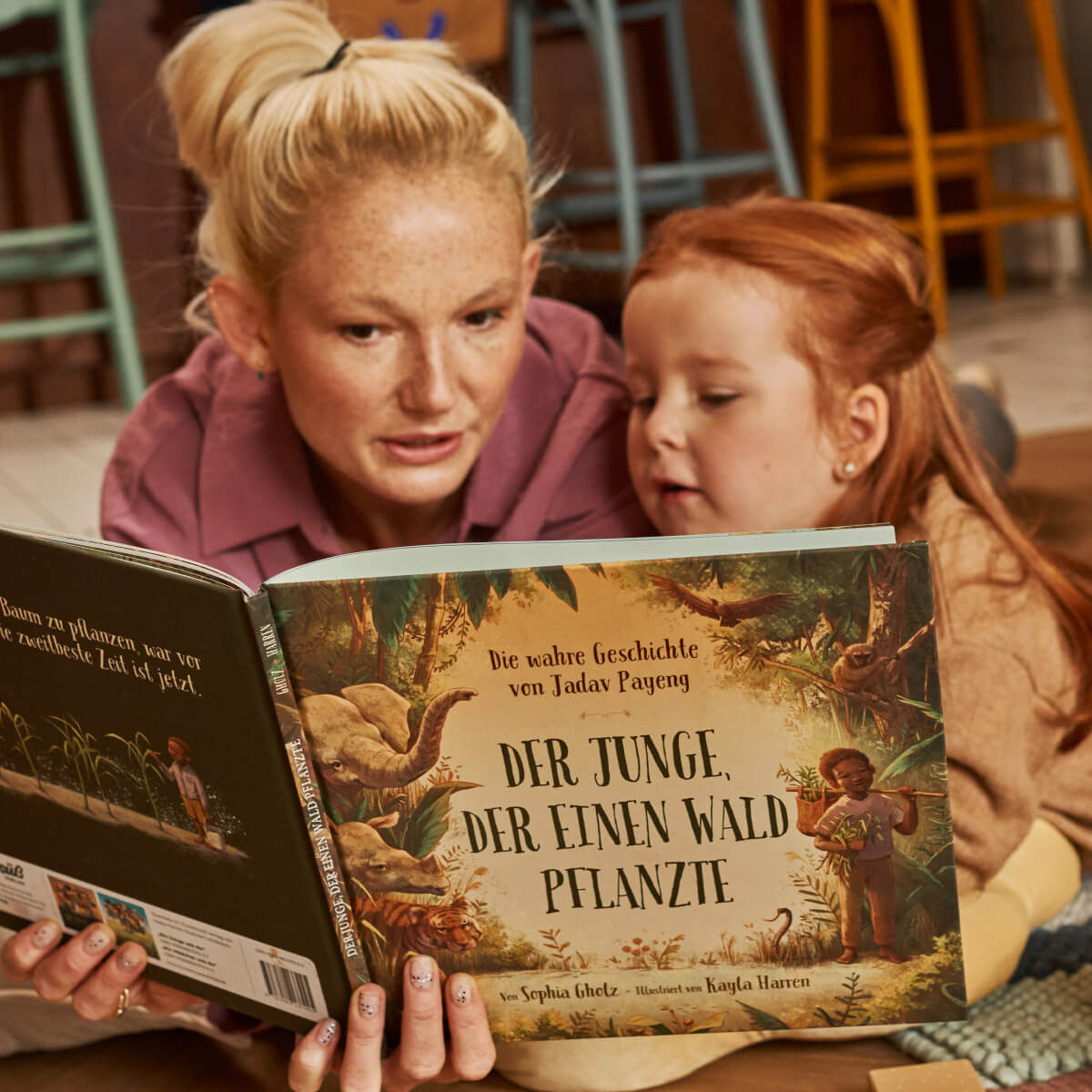 Eine erwachsene Person und ein junges Kind sitzen eng beieinander und lesen "Der Junge, der einen Wald pflanzte" von Sophia Gholz, auf dessen Titelbild Dschungeltiere aus dem Regenwald und ein Kind abgebildet sind.