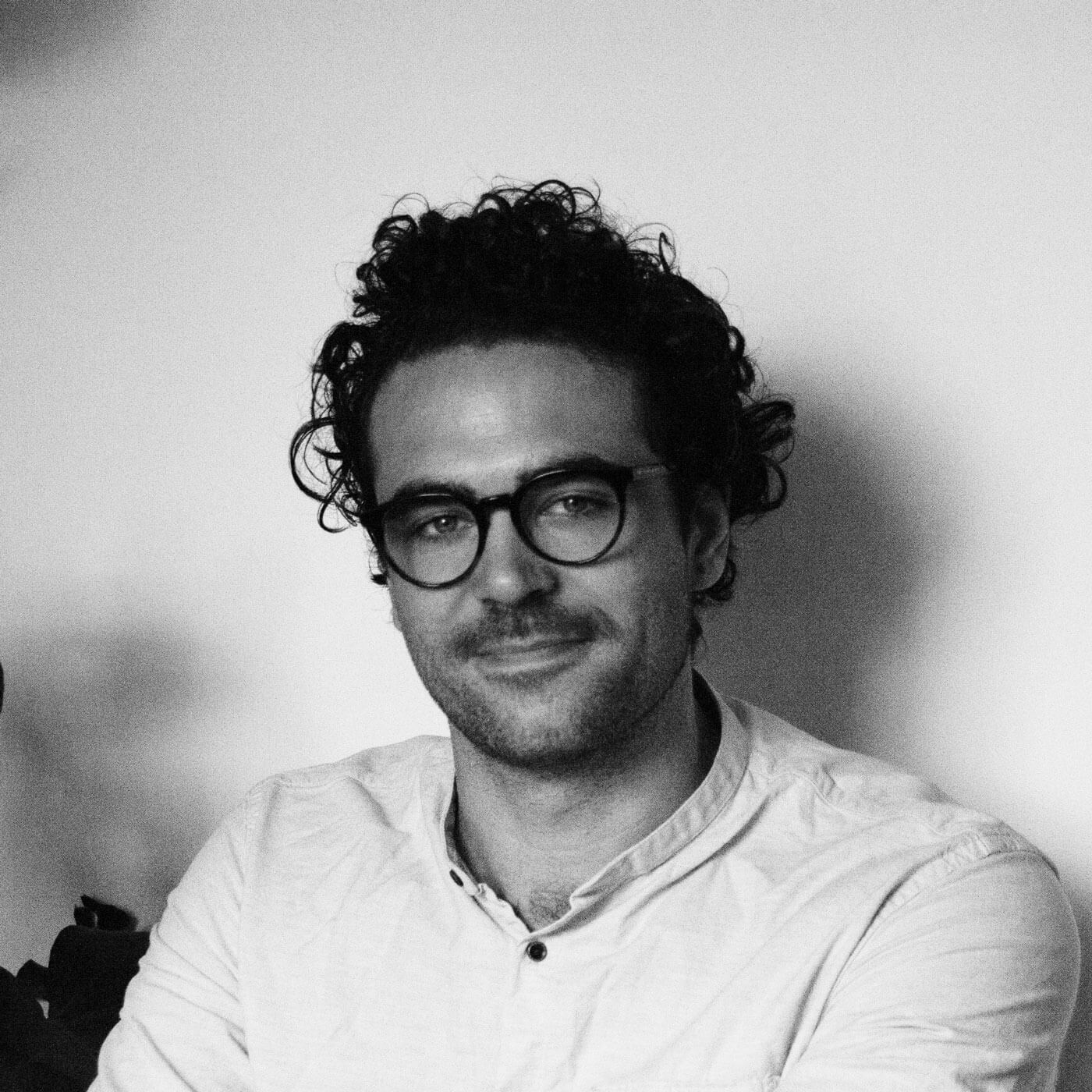 Schwarz-Weiß-Foto des Autors Jonty Howley, der mit lockigem Haar und runder Brille in einem hellen Hemd sitzt und sanft in die Kamera lächelt.