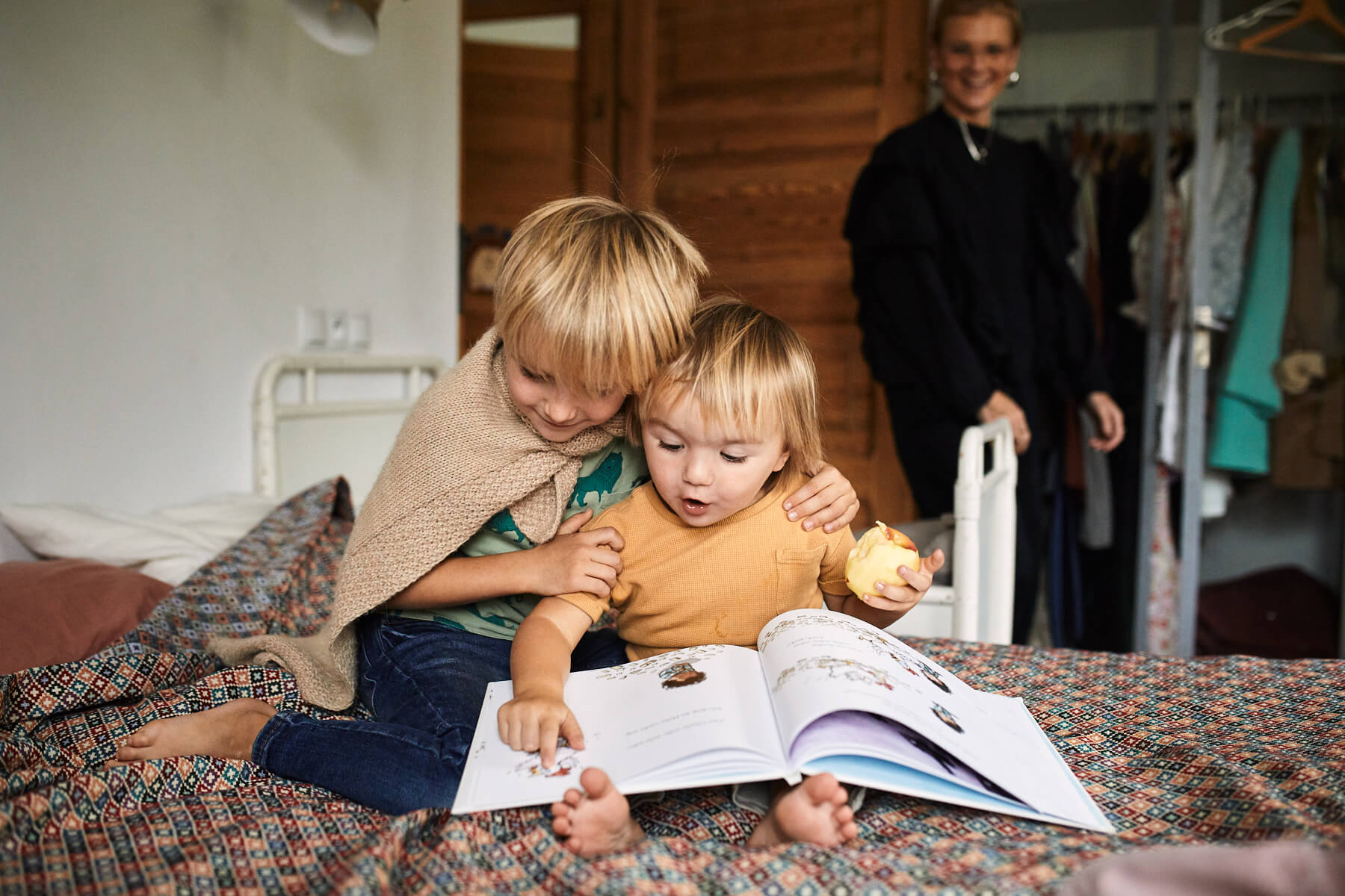 Zwei kleine Kinder sitzen auf einem Bett und schauen sich gemeinsam ein Bilderbuch an. Ein Kind hat einen Arm um das andere gelegt. Ein Erwachsener lächelt im Hintergrund in der Nähe eines offenen Schranks.