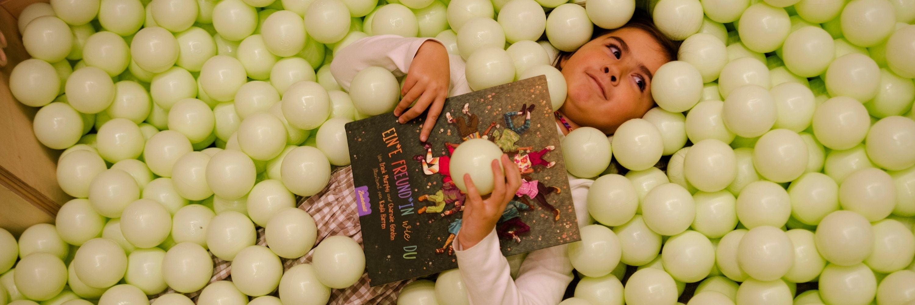 Ein Kind liegt in einer mit blassgrünen Bällen gefüllten Grube, hält ein Buch in der Hand und schaut lächelnd nach oben.