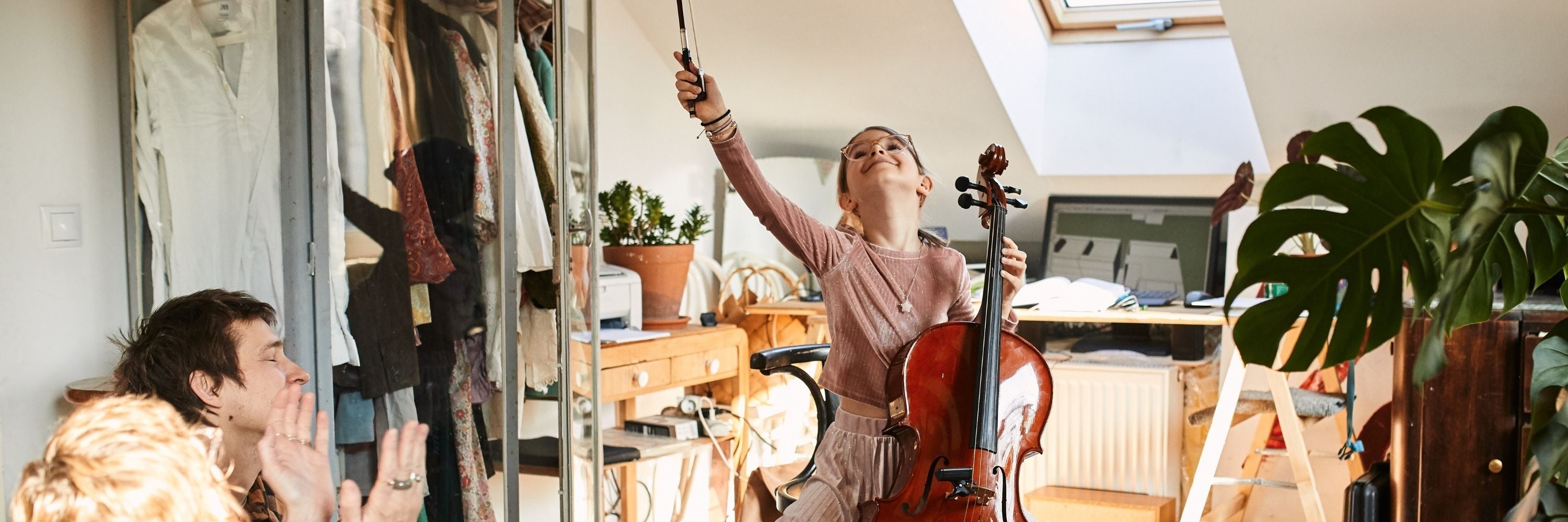 Ein junges Mädchen spielt in einem hellen Raum begeistert Cello und hebt den Bogen in die Luft, während Erwachsene in der Nähe zusehen und klatschen. Der Raum ist gemütlich mit Pflanzen, Kleidern und einfallendem Sonnenlicht.