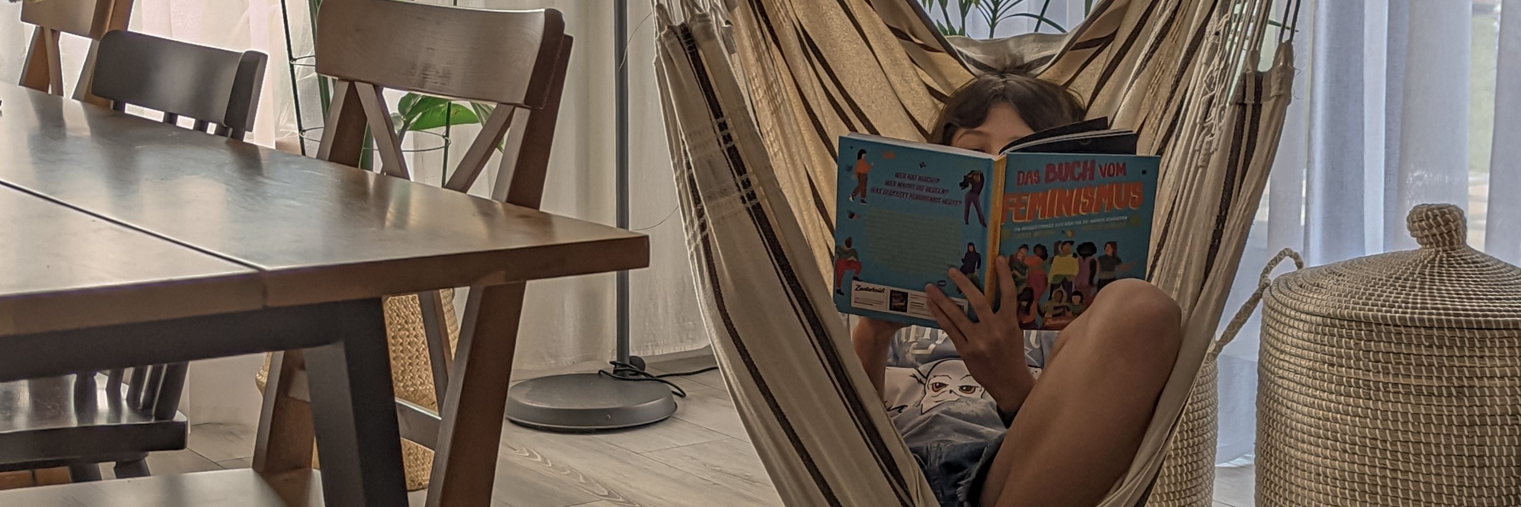 Ein Kind entspannt sich in einem Hängesessel im Haus und liest ein illustriertes Buch. Das Sonnenlicht strömt durch die durchsichtigen Vorhänge, in der Nähe stehen ein Holztisch und Stühle.