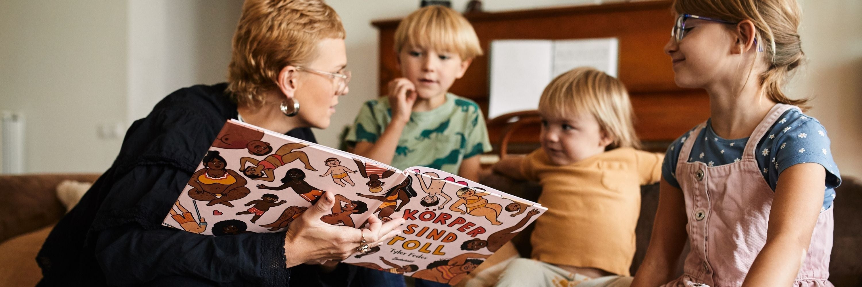 Eine Erwachsene liest drei aufmerksamen Kindern, die um sie herum in einem gemütlichen Raum mit einem Klavier im Hintergrund sitzen, ein buntes Bilderbuch mit dem Titel Körper sind toll vor.