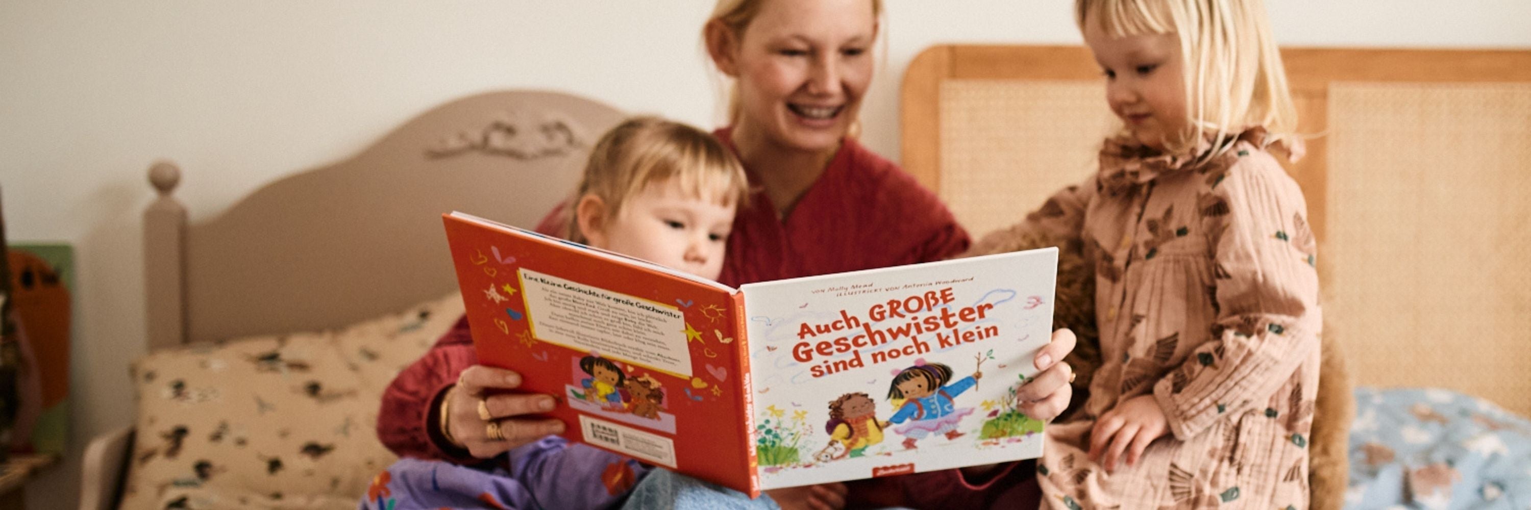 Eine Frau sitzt auf einem Bett und liest zwei jungen Mädchen aus einem deutschen Kinderbuch vor. Ein Mädchen sitzt neben ihr, das andere steht, beide schauen in das Buch und lächeln.
