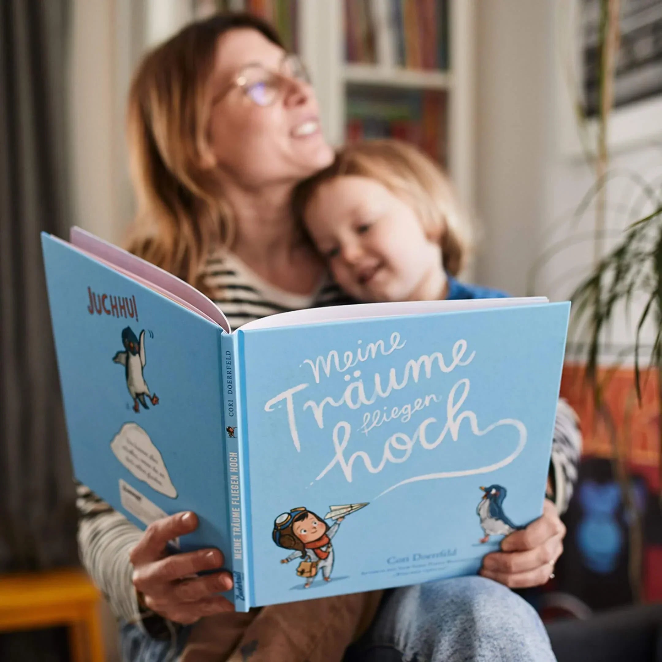 Eine Frau und ein kleines Kind lächeln, während sie ein deutsches Kinderbuch mit dem Titel Meine Träume fliegen hoch lesen. Der blaue Einband zeigt einen Pinguin und ein Mädchen mit Fliegerhut.