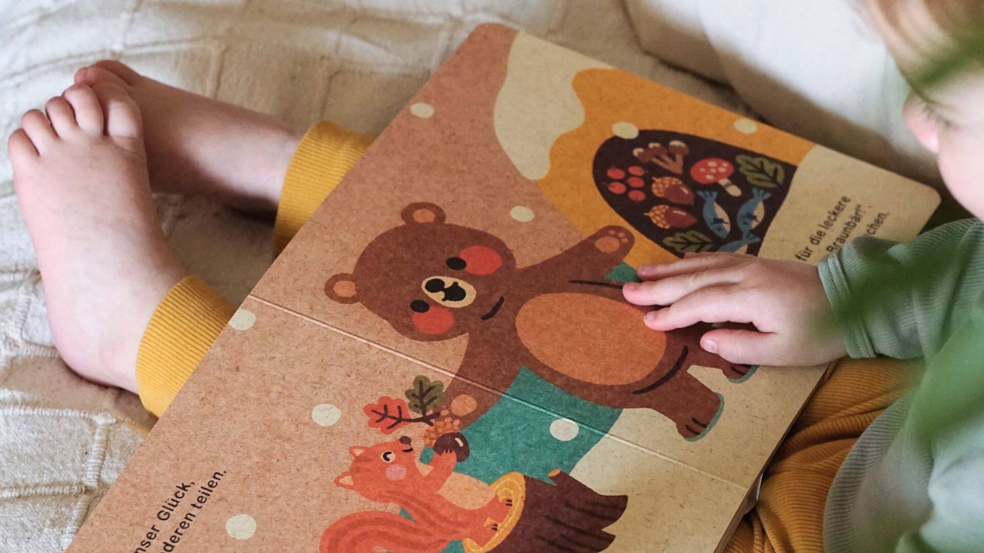 Ein Kind sitzt mit gekreuzten Beinen da und berührt eine Bilderbuchseite aus Pappe, auf der ein lächelnder Bär und ein Eichhörnchen mit einer Eichel abgebildet sind. Das Kind hat eine gelbe Hose und ein grünes Hemd an.