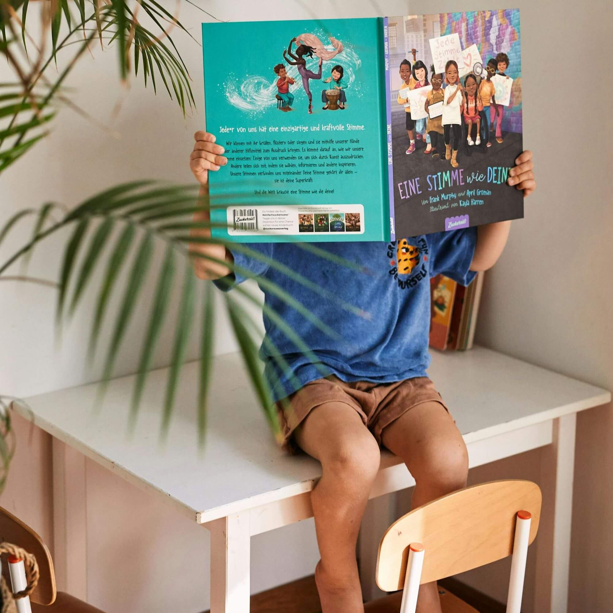 Ein Kind sitzt an einem weißen Schreibtisch und liest "Eine Stimme wie deine" von Frank Murphy und April Groman, wobei sein Gesicht von dem Bilderbuch verdeckt wird. Im Vordergrund erscheint eine grüne Blattpflanze, die einen Moment der Entdeckung einfängt.
