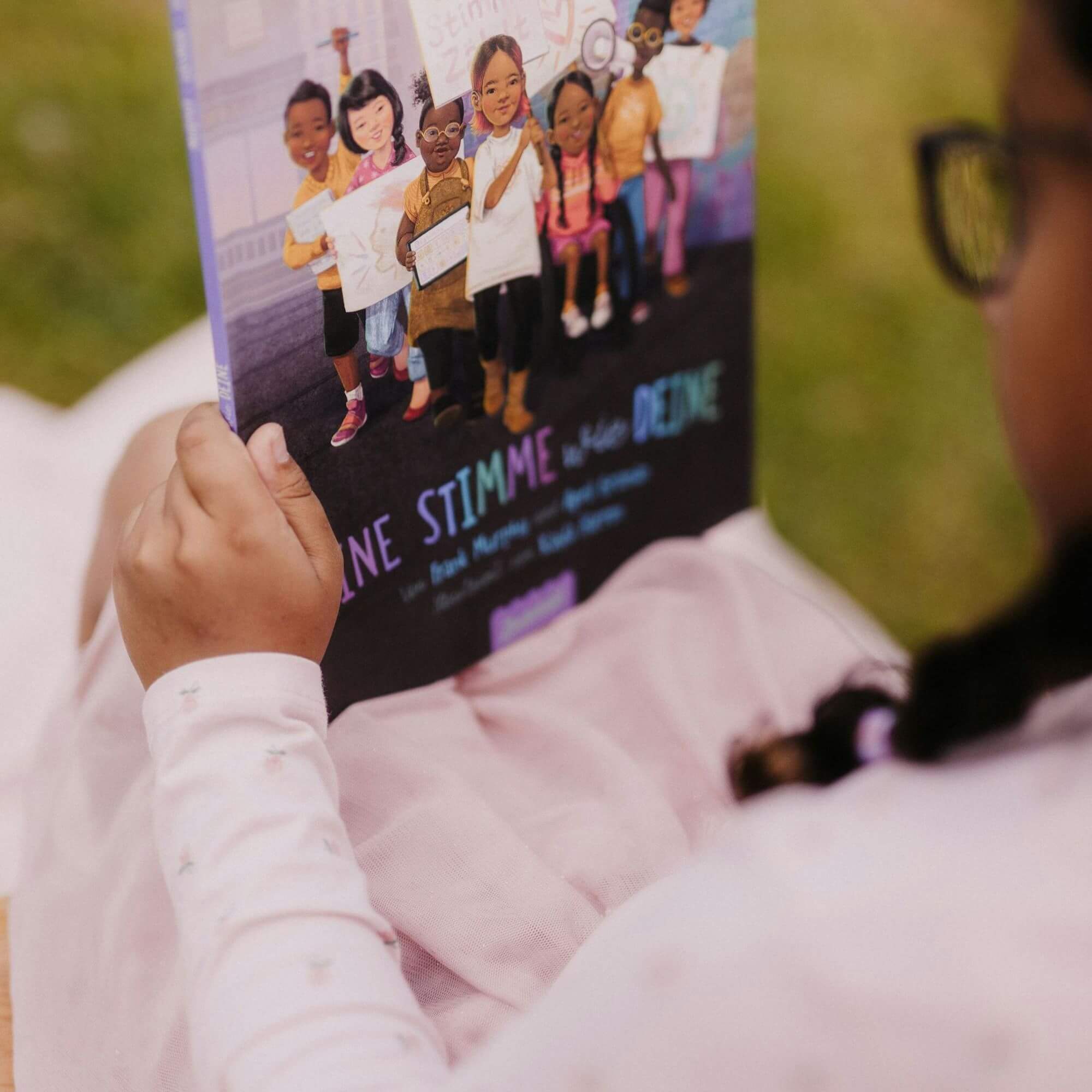 Ein Kind mit Brille und einem hellrosa Kleid sitzt im Freien und liest "Eine Stimme wie deine" von Frank Murphy und April Groman, ein farbenfrohes Buch mit Illustrationen verschiedener Kinder auf dem Umschlag.