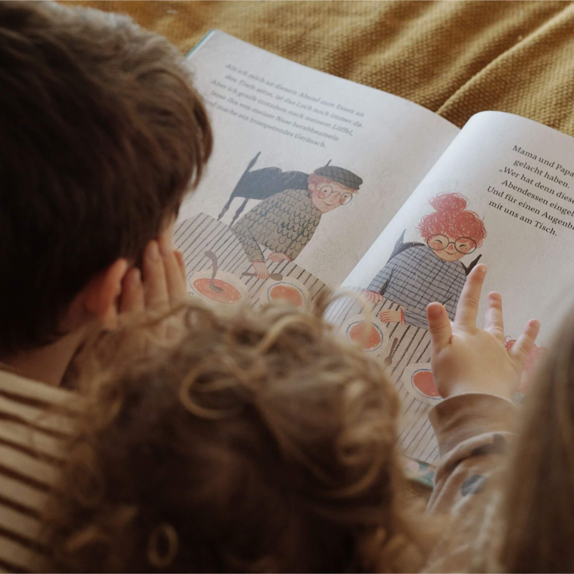 Zwei kleine Kinder sitzen eng beieinander und betrachten ein aufgeschlagenes Exemplar von „Das Loch“ von Lindsay Bonilla. Sie zeigen auf eine Illustration, die zwei ältere Figuren mit Brille an einem Tisch zeigt – vor ihnen stehen mehrere Schüsseln mit Suppe. Der begleitende Text beschreibt eine ruhige, familiäre Szene. Das Bild verbindet Geborgenheit, Erinnerung und die heilende Kraft des Zusammenseins.