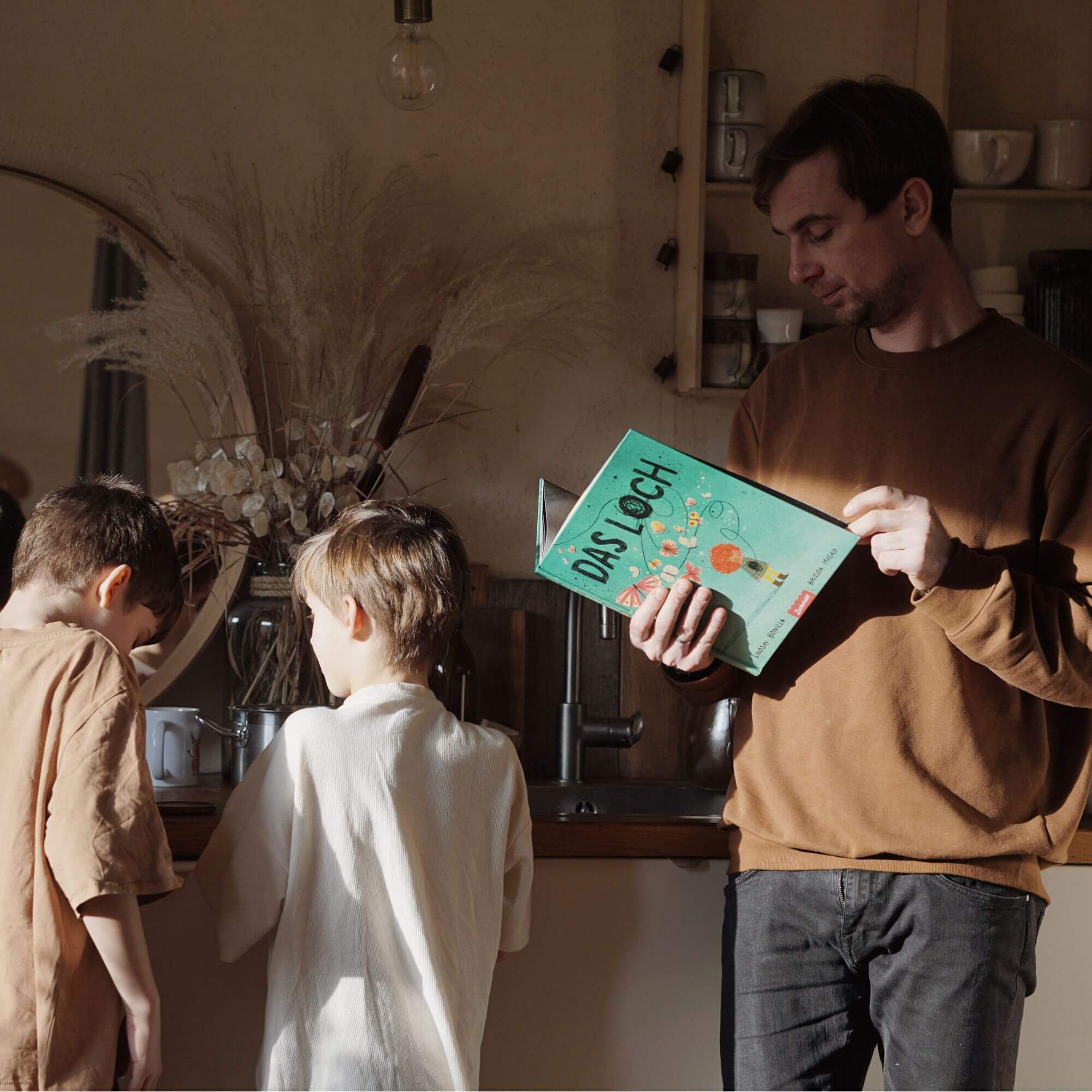 Eine erwachsene Person in einem braunen Pullover hält ein aufgeschlagenes Exemplar von „Das Loch" von Lindsay Bonilla in der Hand, während zwei Kinder in der Nähe an einem sonnenbeschienenen Küchentisch stehen, der mit getrockneten Pflanzen geschmückt ist, die ein Gefühl von Heilung und Erinnerung hervorrufen.
