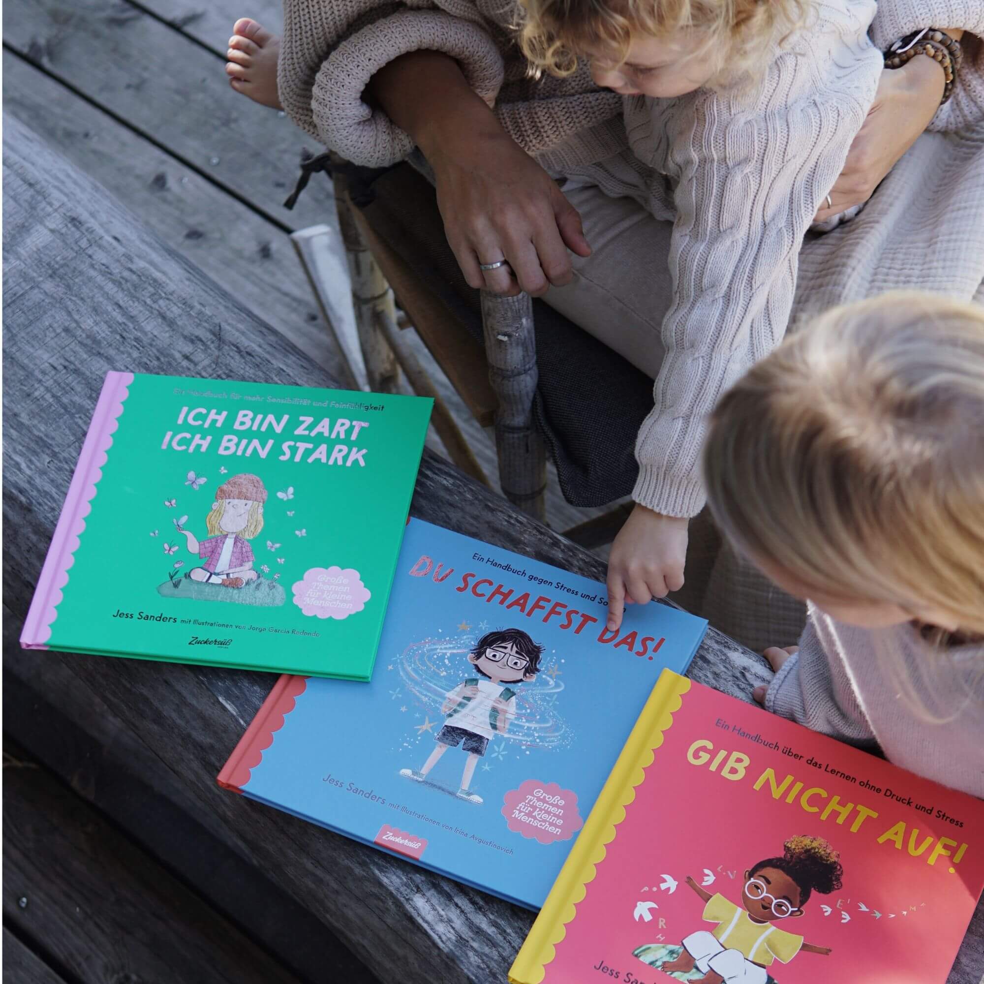 Eine Frau und ein Kind sitzen auf einer Holzbank und betrachten drei bunte Kinderbücher aus dem Mutmach-Paket des Zuckersüß Verlags. Das Kind zeigt auf das blaue Buch, das neben einem rosafarbenen und einem grünen Buch liegt. Die Themen darin behandeln innere Stärke. 
