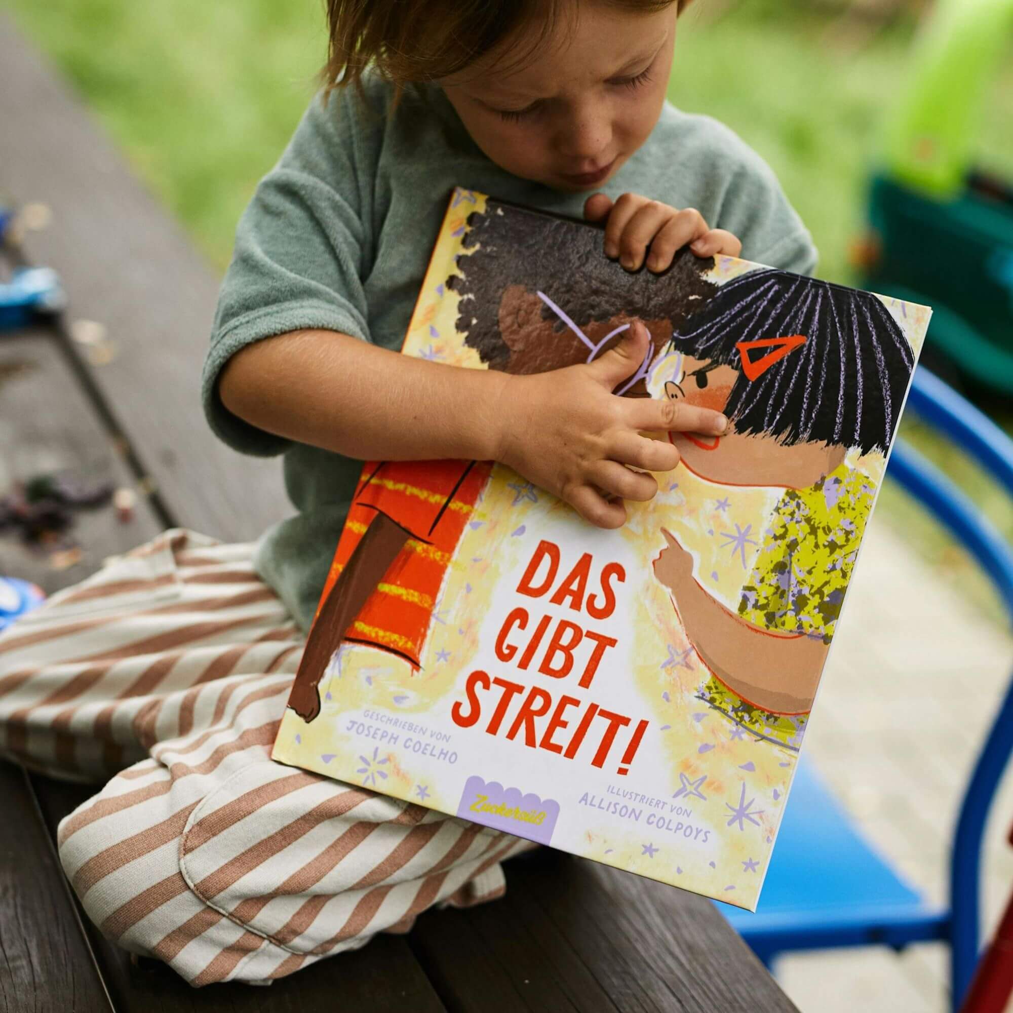 Ein kleines Kind mit gestreifter Hose und grünem Shirt sitzt auf einer Bank und betrachtet neugierig das Bilderbuch „Das gibt Streit!“, dessen Cover zwei gezeichnete Kinder im Streit zeigt.