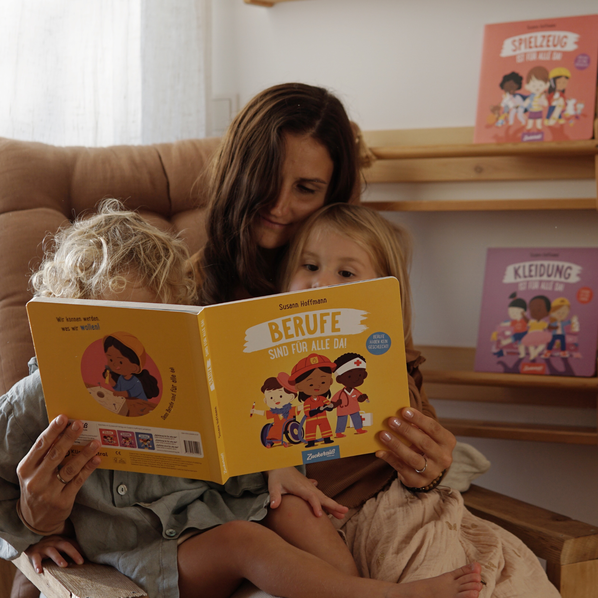Eine Frau liest zwei Kindern auf ihrem Schoß "Freiheit ist für alle da - die komplette Reihe", von Susann Hoffmann, aus dem Zuckersüß Verlag vor. Im Hintergrund stehen bunte Bücher für alle in Regalen.