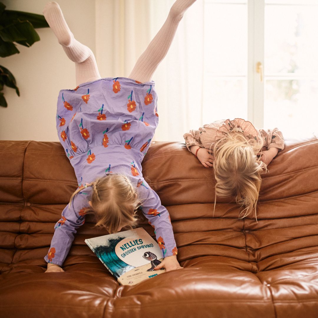 Zwei junge Mädchen sitzen auf einem braunen Sofa; die eine steht auf dem Kopf und liest "Nellies großer Sprung" aus dem Zuckersüß Verlag, der Mut und innere Stärke hervorhebt, während die andere sich mit gesenktem Kopf über die Lehne lehnt.