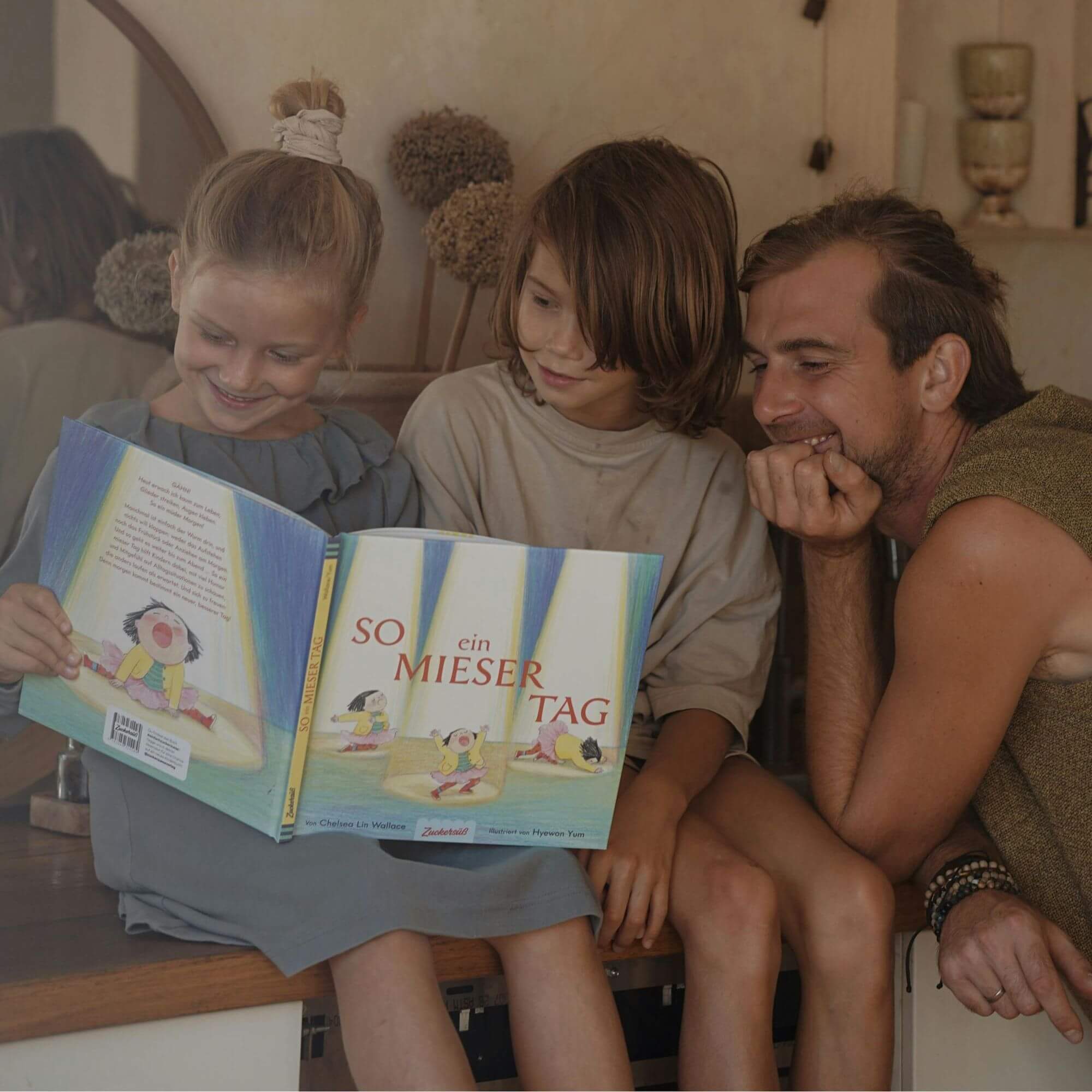 Ein lächelnder Mann sitzt neben zwei Kindern auf einem Tresen und liest fröhlich "So ein mieser Tag" von Chelsea Lin Wallace, während warmes Licht den gemütlichen Raum erfüllt.