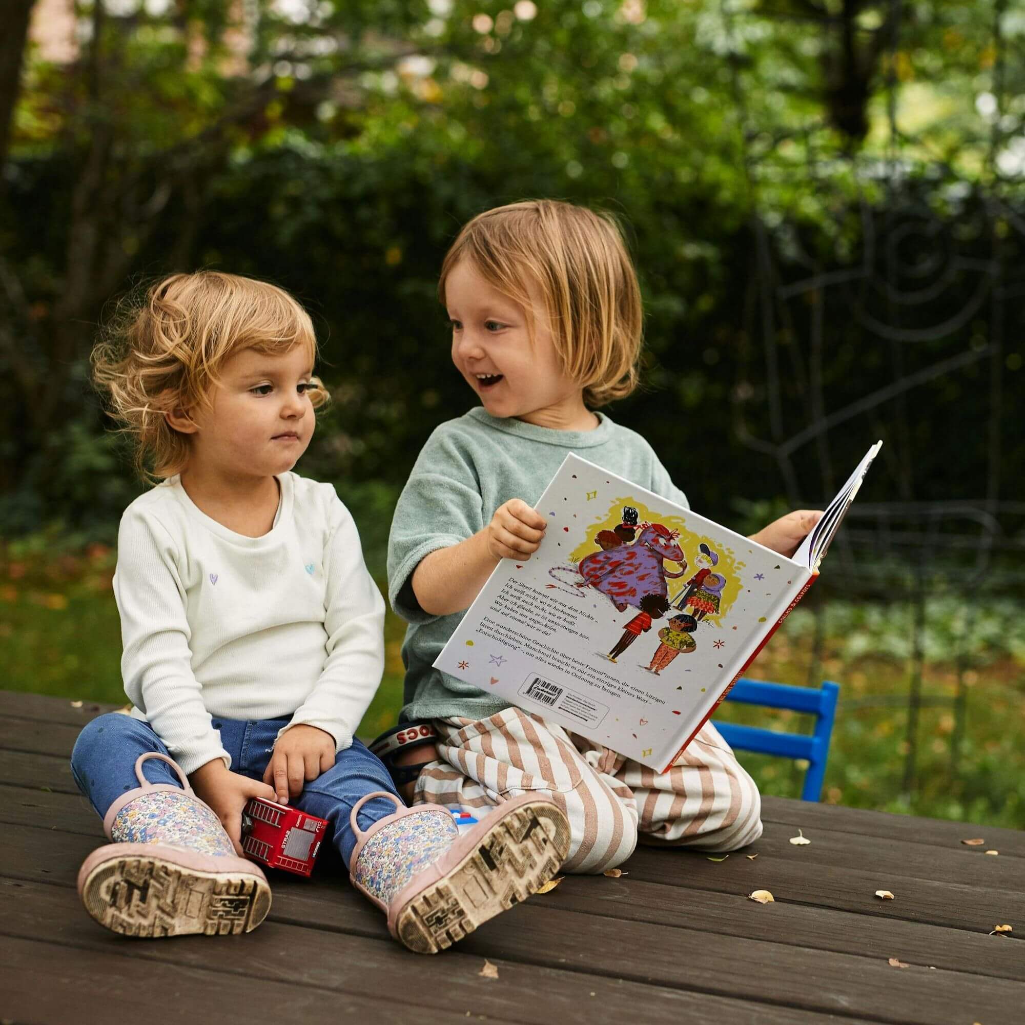 Zwei Kleinkinder sitzen draußen an einem Holztisch. Eines von ihnen hält lächelnd das Bilderbuch "Das gibt Streit!" von Joseph Coelho und Allison Colpoys, während das andere ein Spielzeugauto in der Hand hält. Im Hintergrund ist grüne Vegetation zu sehen.