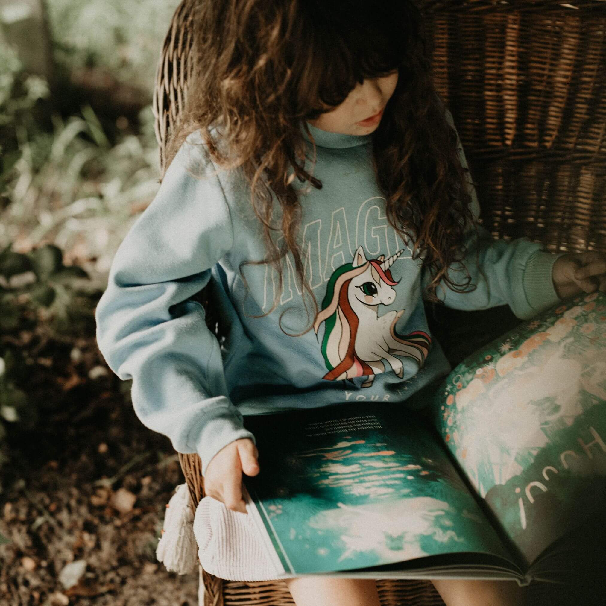 Ein kleines Kind mit langen braunen Haaren sitzt in einem Korbstuhl im Freien, trägt ein blaues Sweatshirt mit einer Einhörner-Grafik und liest "Matteo glaubt an Einhörner" von Jackie Azúa Kramer & Jonah Kramer, das mit bunten Illustrationen versehen ist.