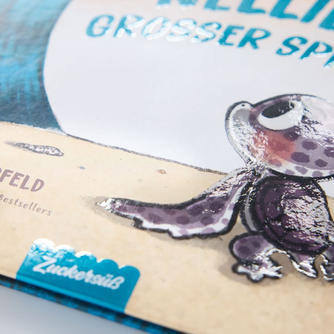 Nahaufnahme des Covers von "Nellies großer Sprung" aus dem Zuckersüß Verlag: eine illustrierte Schildkröte mit großen Augen und lila Flecken auf Sand, ein blaues Zuckersüß-Label und glänzende Prägedetails - eine Geschichte über Mut und innere Stärke.