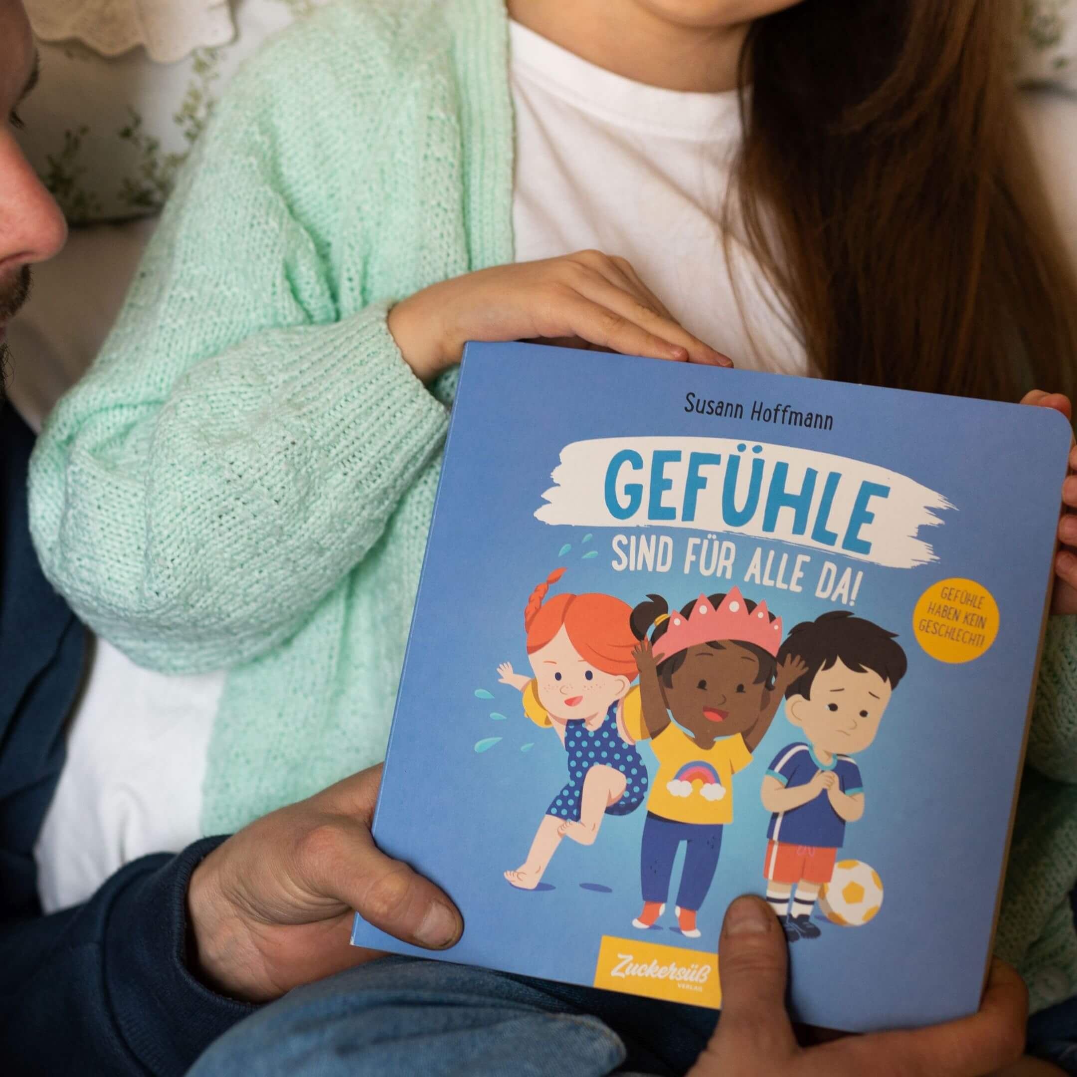 Ein Kind hält das Bilderbuch „Gefühle sind für alle da!“ von Susann Hoffmann, ein Erwachsener stützt es von unten. Auf dem Cover sind drei Kinder mit unterschiedlichen Emotionen zu sehen.