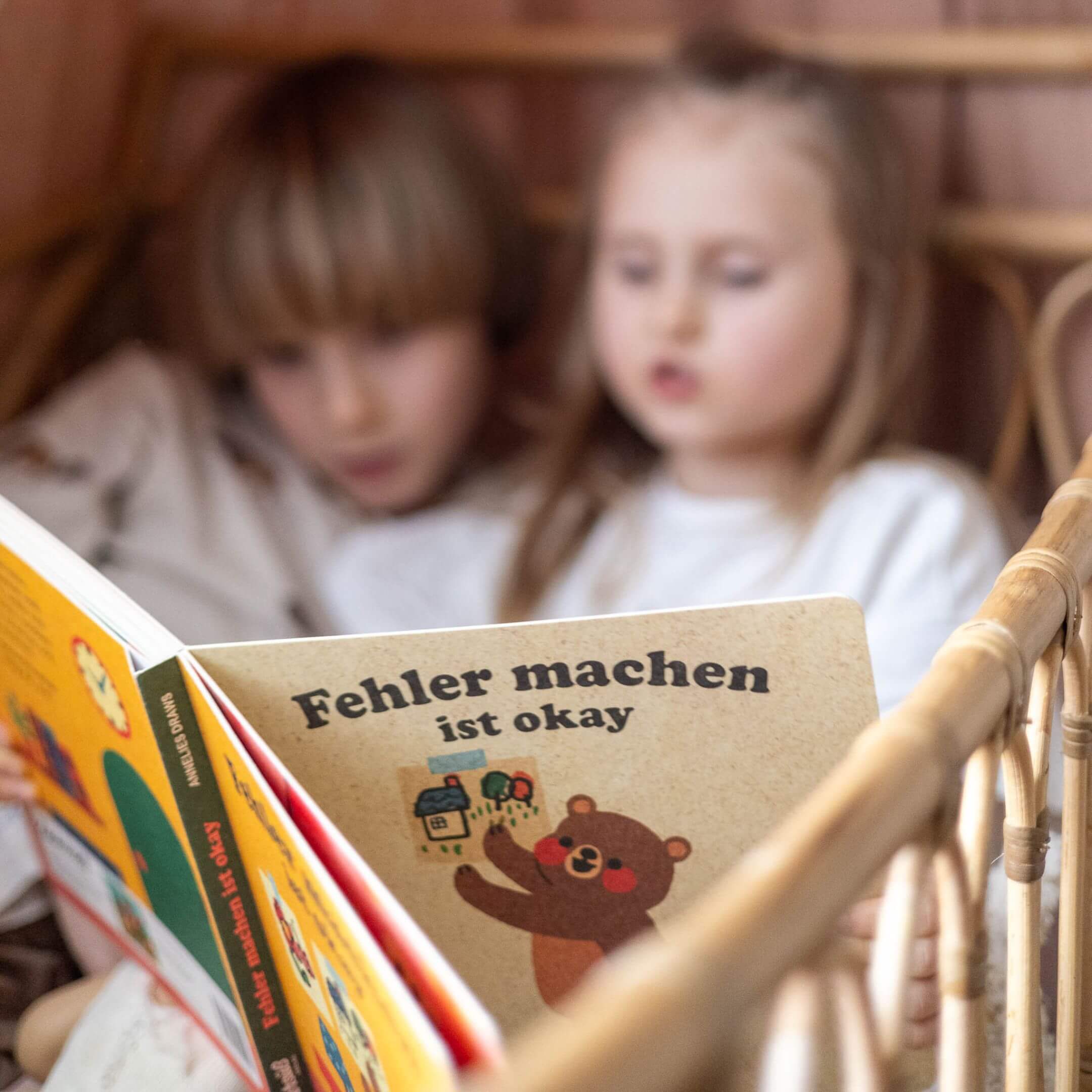 Zwei Kinder sitzen zusammen in einem Korbsessel und lesen "Fehler machen ist ok" von Annelies Draws. Das illustrierte Buch zeigt einen Bären im Retrostil, und die Kinder sind ganz auf die Geschichte konzentriert.