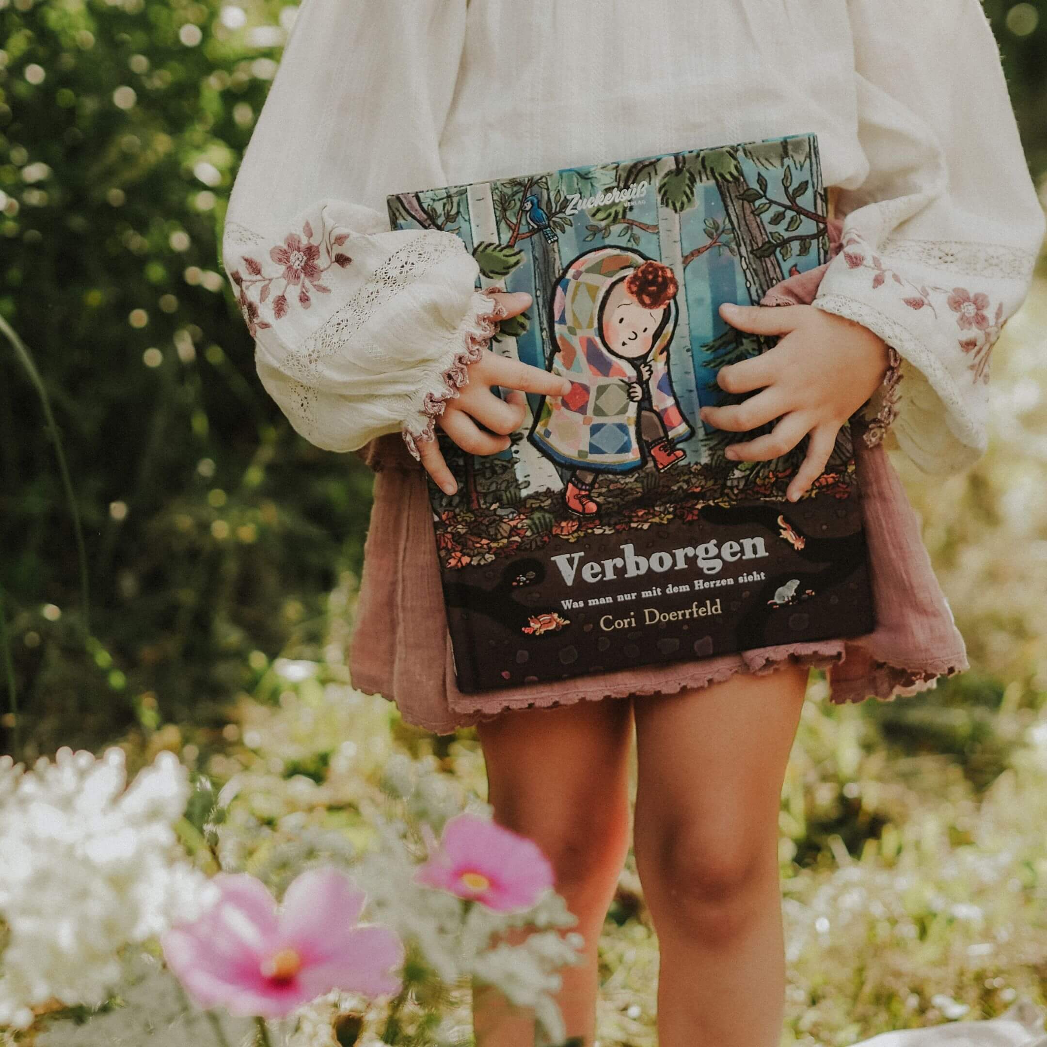 Ein Kind in einer weißen bestickten Bluse und einem rosa Rock steht draußen zwischen grünem Gras und rosa Blumen und hält das Buch "Verborgen" aus dem „Best of Cori Doerrfeld“ Paket vom Zuckersüß Verlag in der Hand.