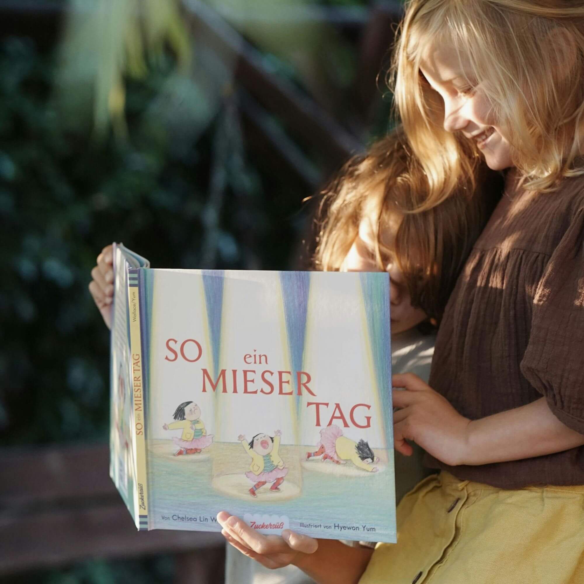 Zwei lächelnde Kinder lesen "So ein mieser Tag" von Chelsea Lin Wallace draußen im sanften Sonnenlicht. Das Cover zeigt Cartoonfiguren, die spielerisch im Wasser planschen.