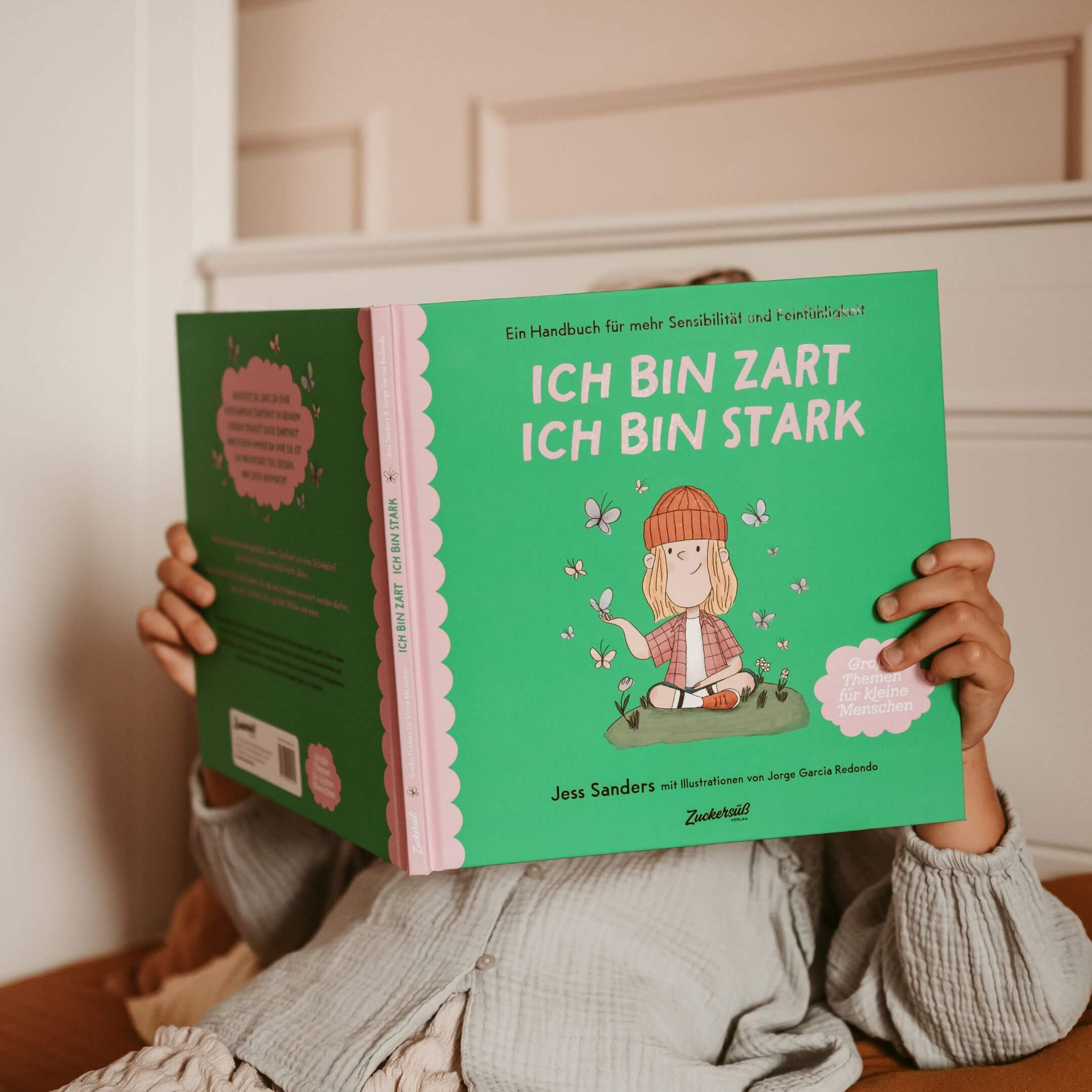 Eine Person in einem hellen Schlafanzug sitzt und hält das Buch von Jessica Sanders "Ich bin zart. Ich bin stark." Das Cover zeigt die Illustration eines Kindes, das von Blumen und Blättern umgeben ist.