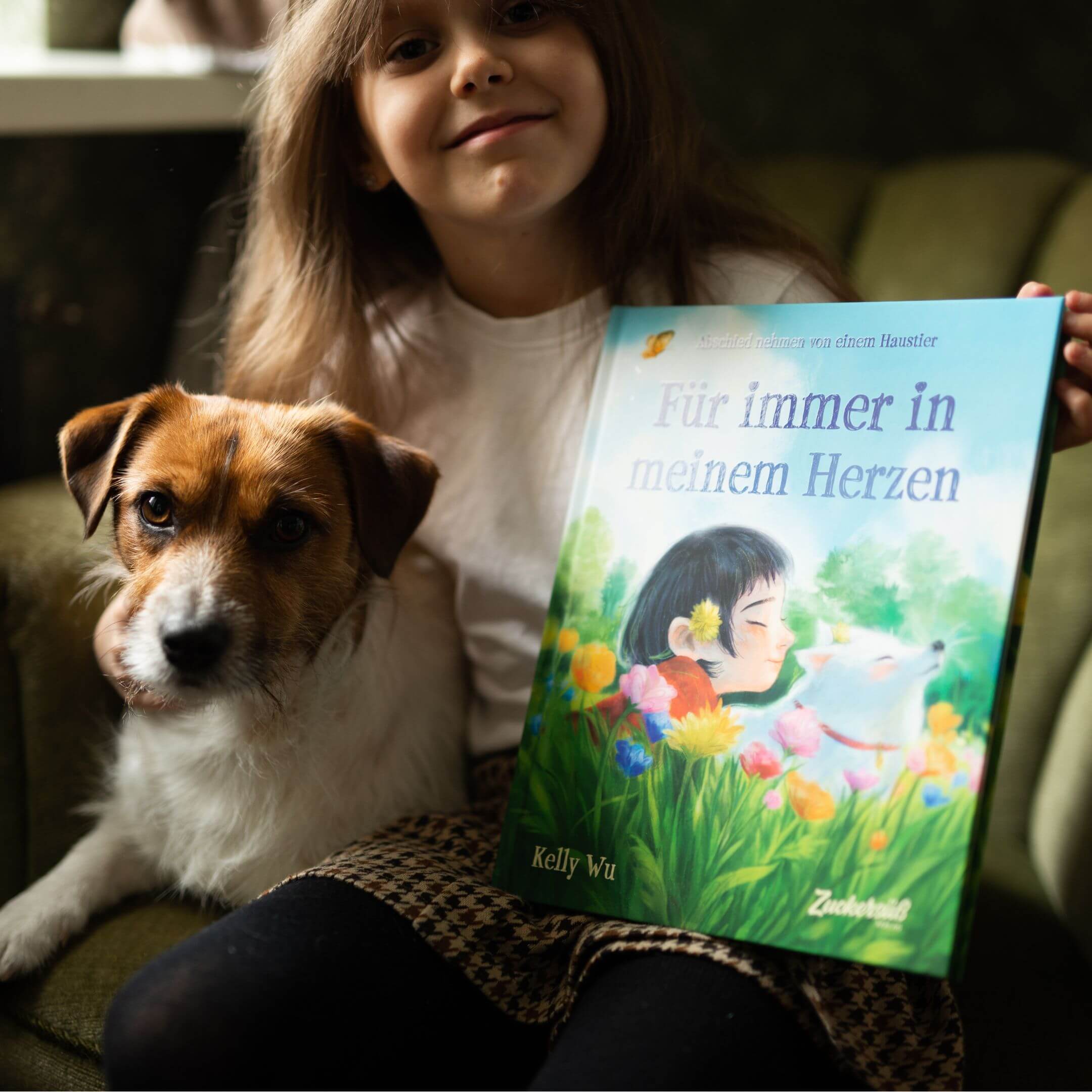Ein lächelndes junges Kind sitzt auf einem grünen Stuhl, hält einen kleinen braun-weißen Hund und "Für immer in meinem Herzen" von Kelly Wu - ein Kinderbuch über Trauer, dessen Cover ein Mädchen inmitten von Blumen und Tieren zeigt.