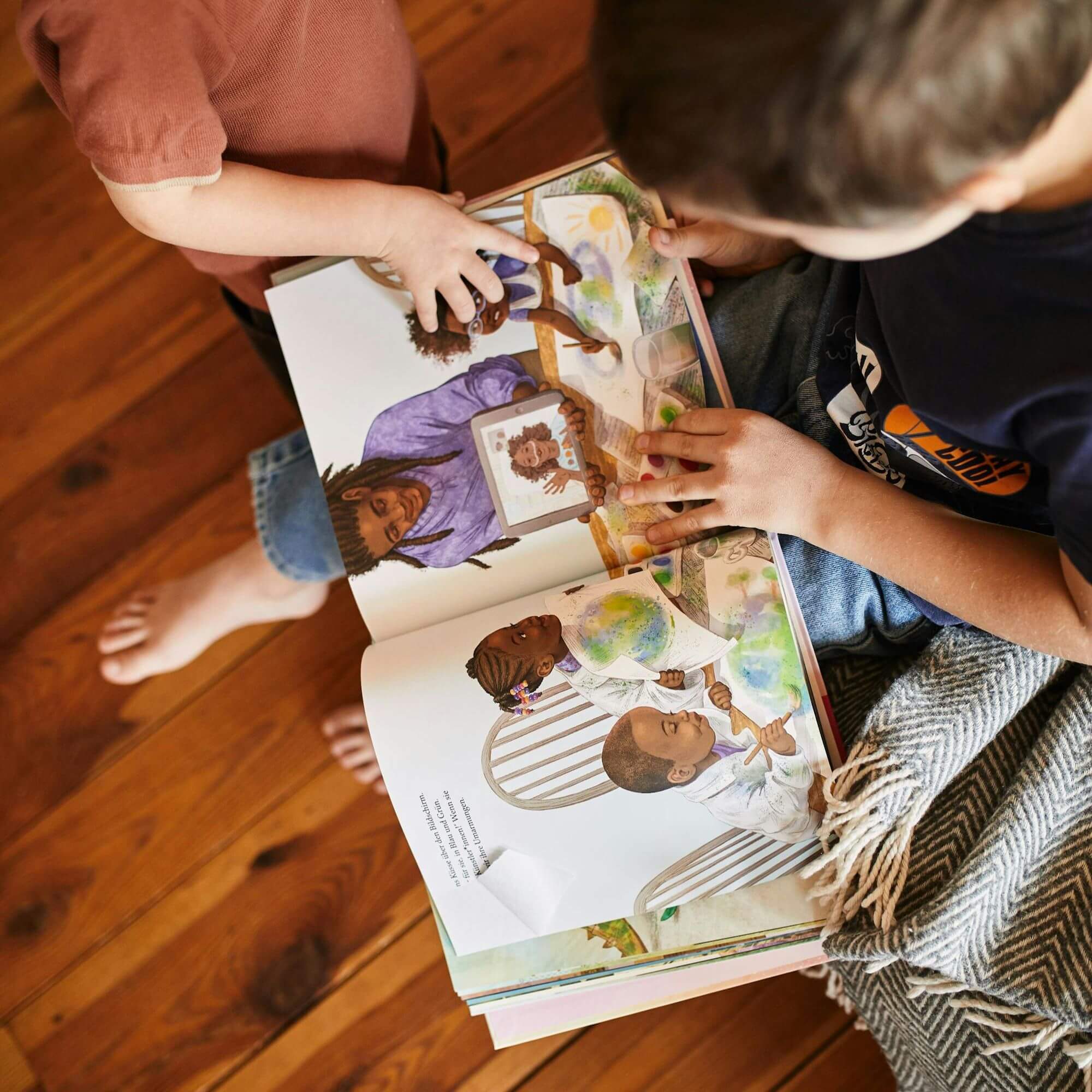 Zwei Kinder sitzen auf einer Decke auf einem Holzboden und blättern gemeinsam im Buch „Alle spielen mit“ von Caroline Cupp. Die Doppelseite zeigt diverse Kinder beim Lesen und Spielen, darunter auch ein Kind im Rollstuhl. Die farbenfrohen Illustrationen feiern Vielfalt, Gemeinschaft und Neugier.