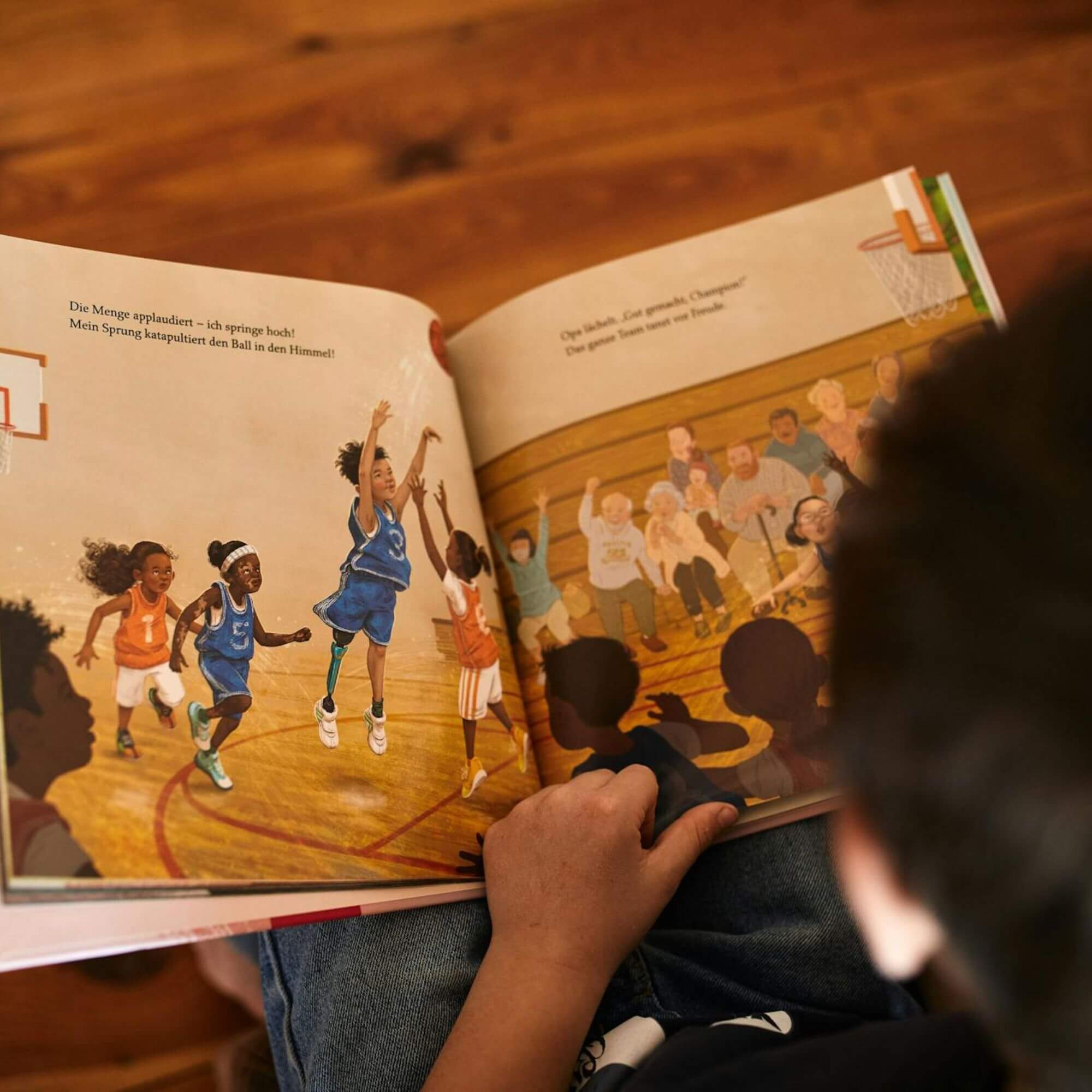 Ein Kind liest "Alle spielen mit" von Caroline Cupp, ein Bilderbuch mit Text, das auf einer Seite verschiedene Kinder beim Basketballspiel und auf der anderen Seite eine jubelnde Menge in der Turnhalle zeigt, die das aufgeschlagene Buch in den Händen hält.