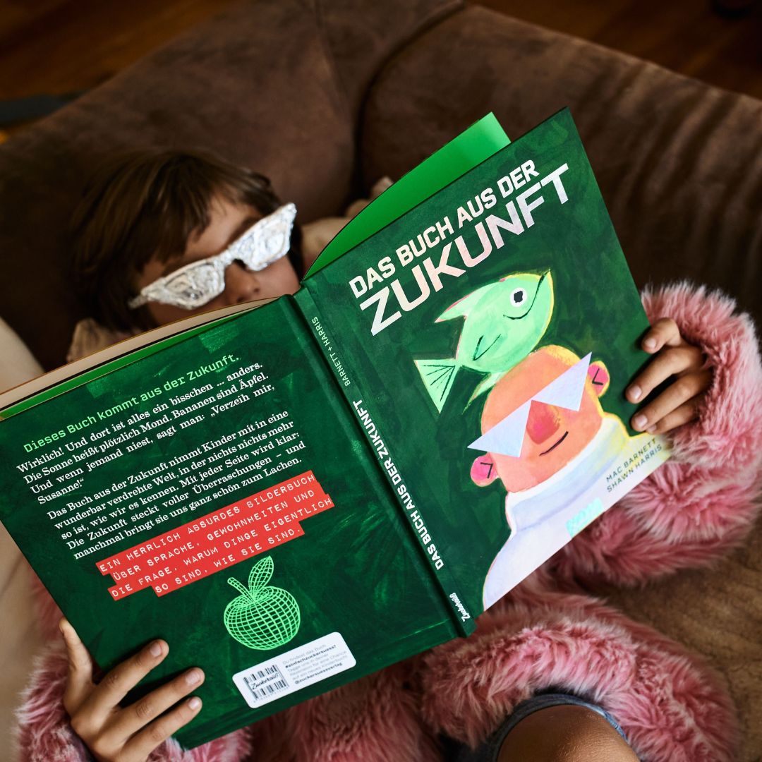 Eine Person mit einer glitzernden Brille und einer rosa Pelzjacke liegt auf einem Sofa und liest „Das Buch aus der Zukunft“, ein außergewöhnliches Bilderbuch aus dem Zuckersüß Verlag. Das deutsche Buch hat einen grünen Einband, auf dem ein Fisch und eine Person abgebildet sind.