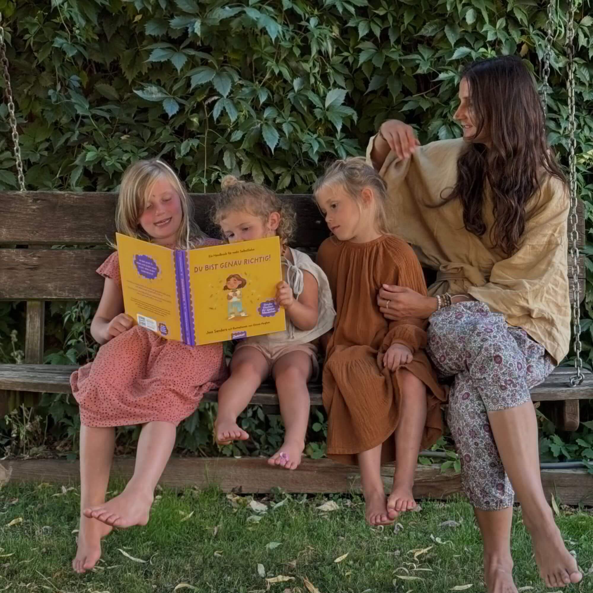 Eine Frau und drei junge Mädchen sitzen barfuß auf einer Holzschaukel im Freien, umgeben von Weinreben. Die Mädchen lesen gemeinsam 