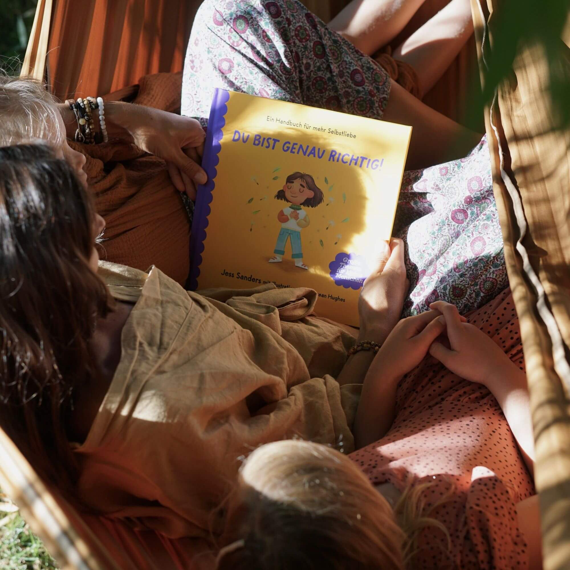 Drei Menschen entspannen sich gemeinsam in einer Hängematte und lesen "Ich bin genau richtig" von Jessica Sander - eine herzerwärmende Geschichte über Selbstliebe - während das sanfte Sonnenlicht durch die Blätter fällt.