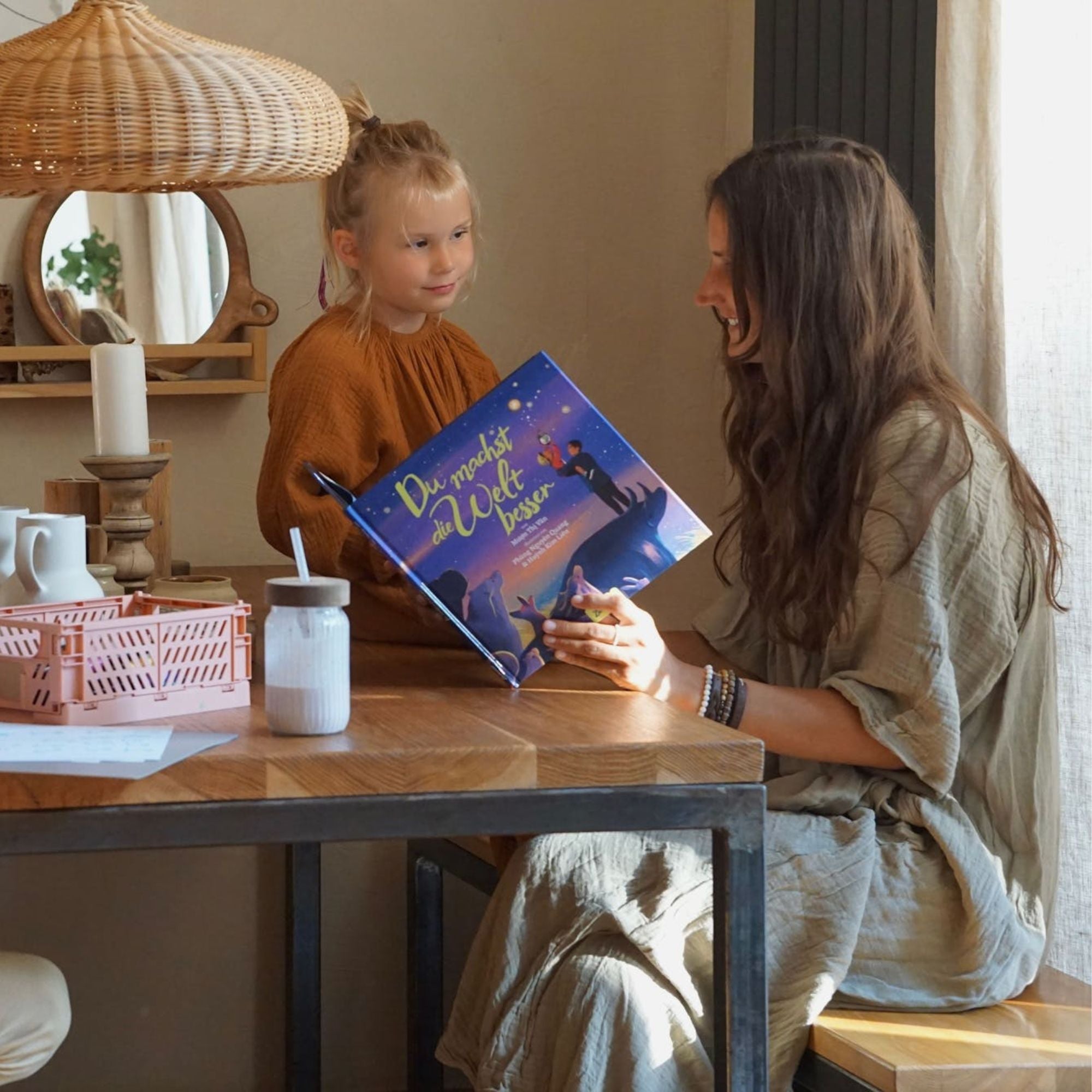 Eine Frau und ein junges Mädchen sitzen an einem Holztisch in einem gemütlichen Zimmer. Lächelnd zeigt die Frau dem Mädchen "Du machst die Welt besser" vom Zuckersüß Verlag, während ein rosa Korb und ein Getränk mit Strohhalm auf dem Tisch stehen.