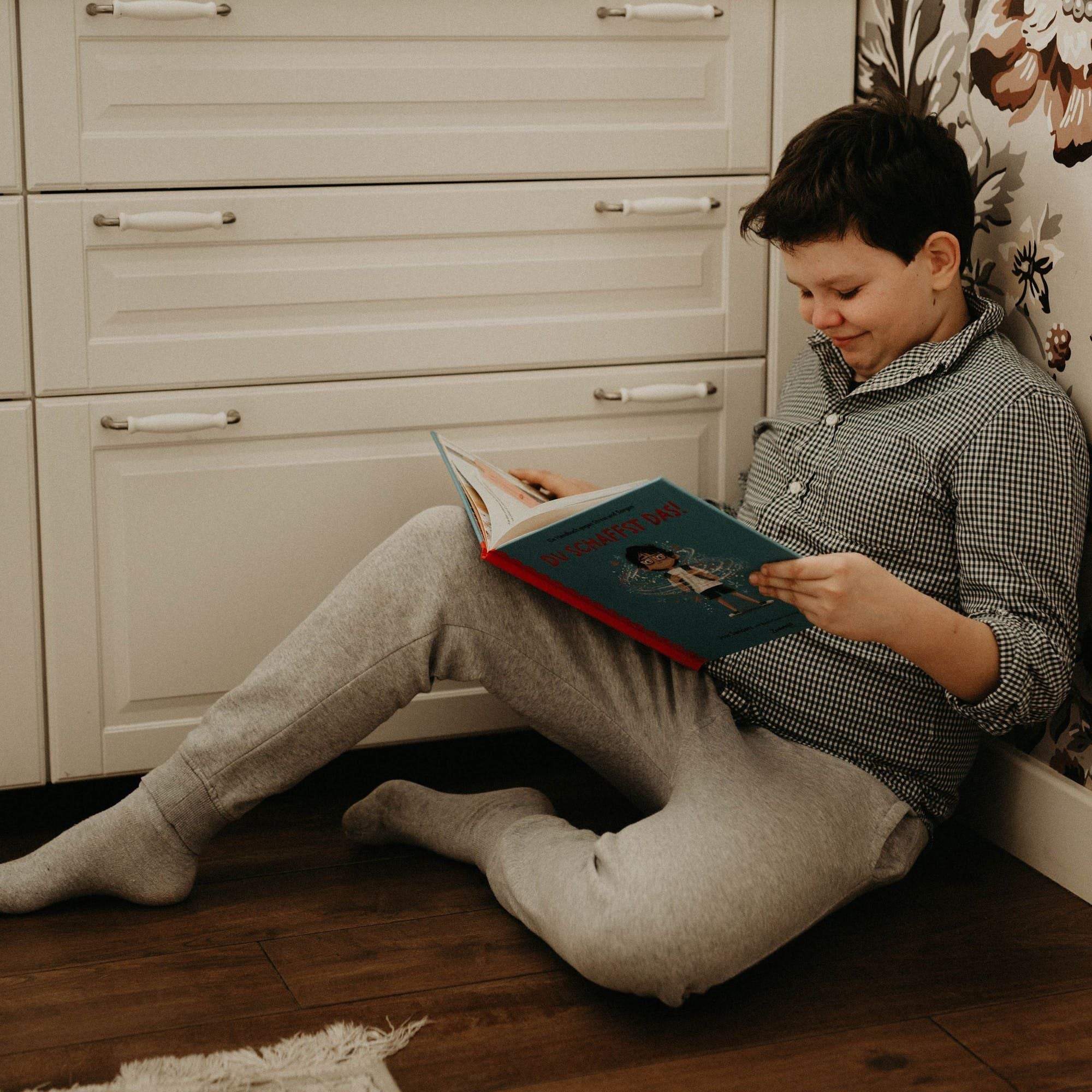 Ein Junge in einem karierten Hemd und einer grauen Jogginghose sitzt auf einem Holzboden zwischen weißen Schränken und liest lächelnd in einem bunten Buch aus seinem School Starter Pack des Zuckersüß Verlags.