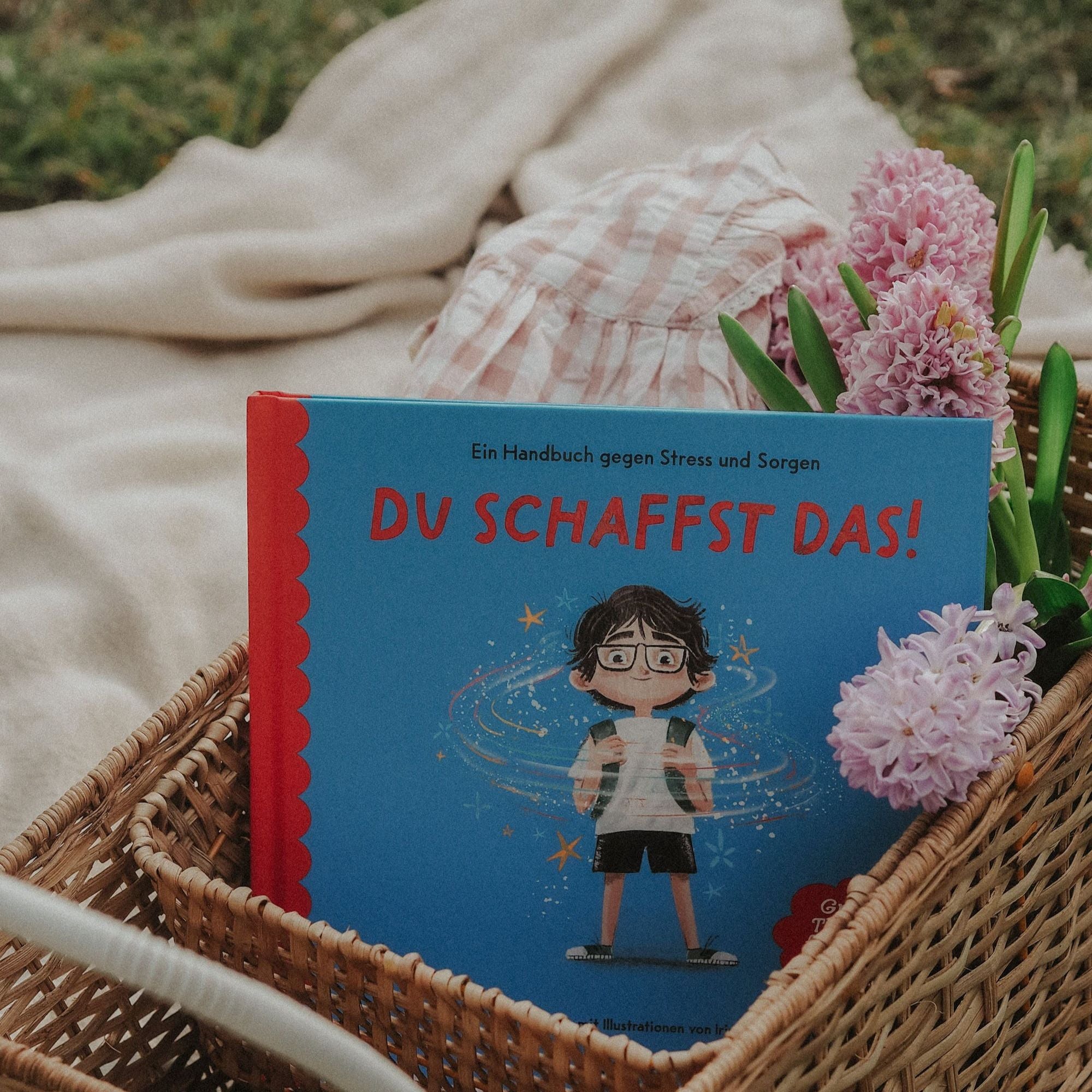 Das "School Starter Pack" des Zuckersüß Verlags mit dem Kinderbuch "Du schaffst das!" zur Förderung von Mut und Selbstvertrauen wird in einem geflochtenen Korb mit rosa Blumen auf einer Outdoor-Decke präsentiert.