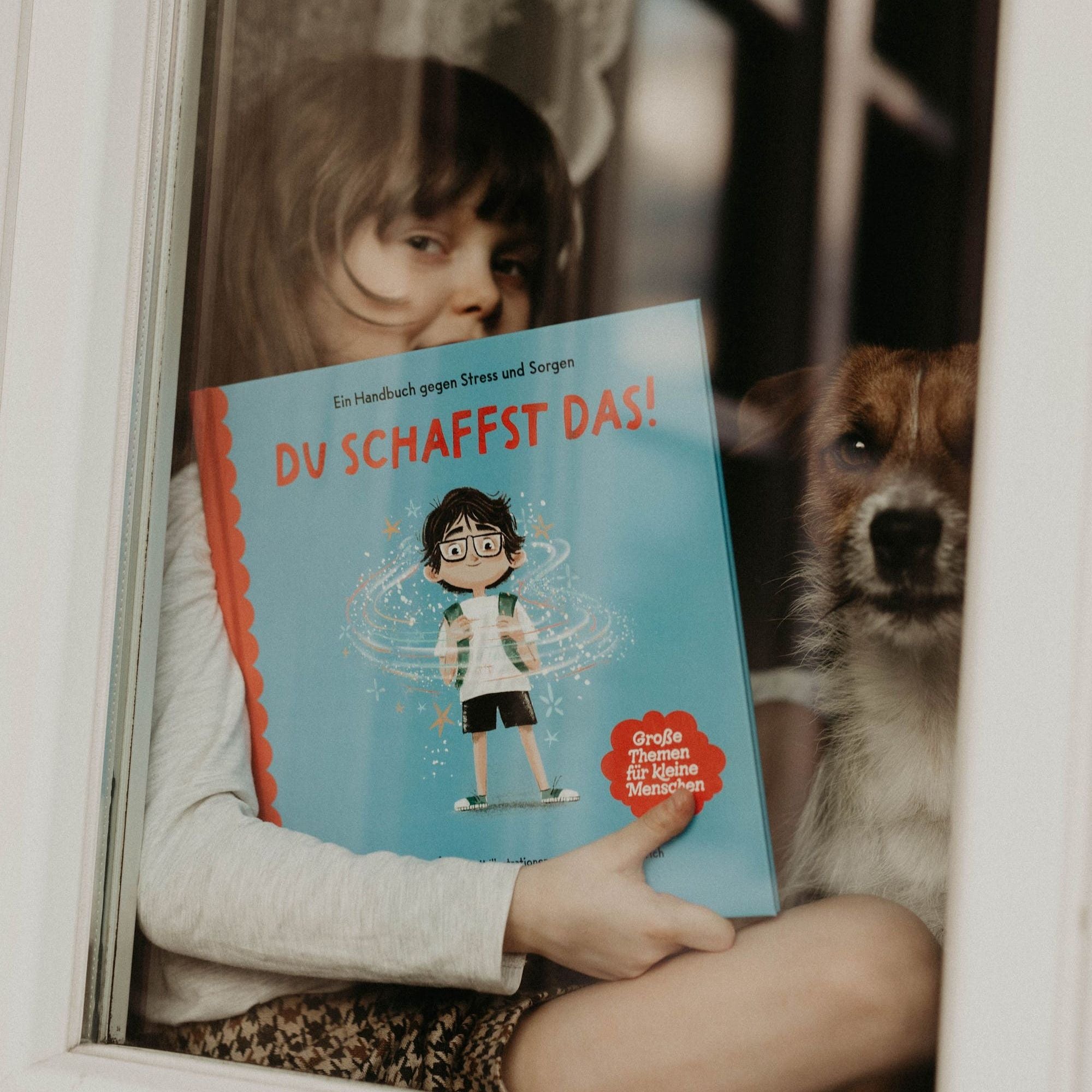 Ein Kind sitzt mit einem Hund am Fenster und hält das Buch „Du schaffst das!“ von Jess Sanders – über Mut und Selbstvertrauen – hoch, das Teil des „School Starter Pack“ des Zuckersüß Verlags ist.