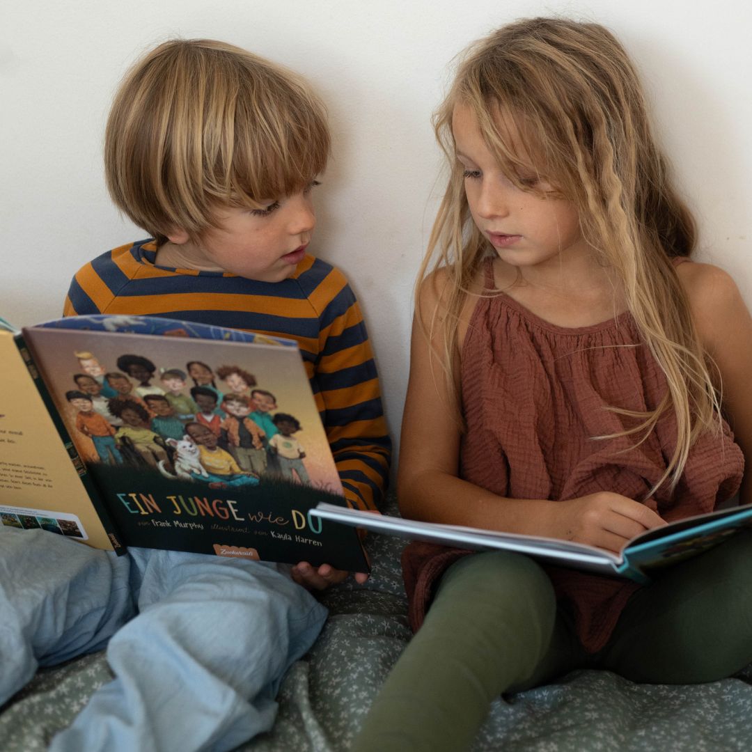 Zwei kleine Kinder sitzen auf einem Bett und lesen Frank Murphys Diversität Kinderbuch, Ein Junge wie Du. Der Junge trägt ein gestreiftes Hemd, das Mädchen das ärmellose Kleid des Zuckersüß Verlags "Eine Welt wie diese - Mädchen-Kollektion", beide mit Blick auf das Buch.