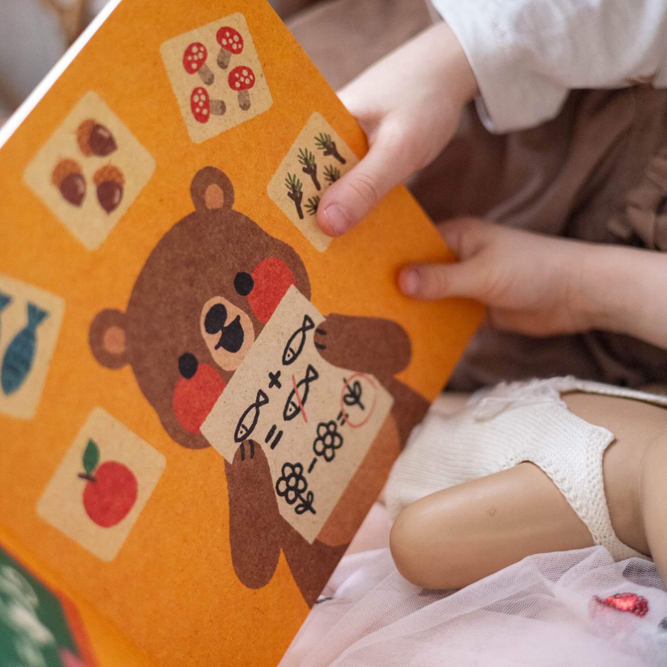 Ein Kind hält ein illustriertes "Fehler machen ist ok"-Kinderbuch von Annelies Draws in der Hand. Auf dem Umschlag ist ein Bär abgebildet, umgeben von Zeichnungen von Lebensmitteln und Pflanzen im Retrostil, mit den Händen des Kindes und einem Teil einer Puppe im Vordergrund.