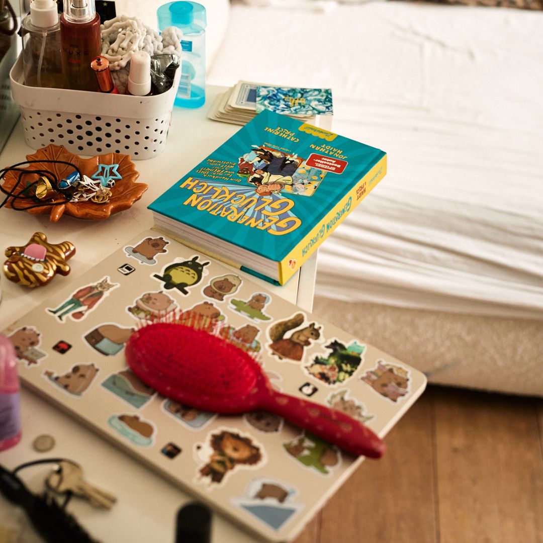 Auf dem unordentlichen Nachttisch stehen ein Korb mit Toilettenartikeln, Schmuck, eine rote Haarbürste, das Buch Generation Glücklich aus dem Zuckersüß Verlag und ein Laptop mit Tieraufklebern - neben einem Bett mit weißer Bettwäsche.